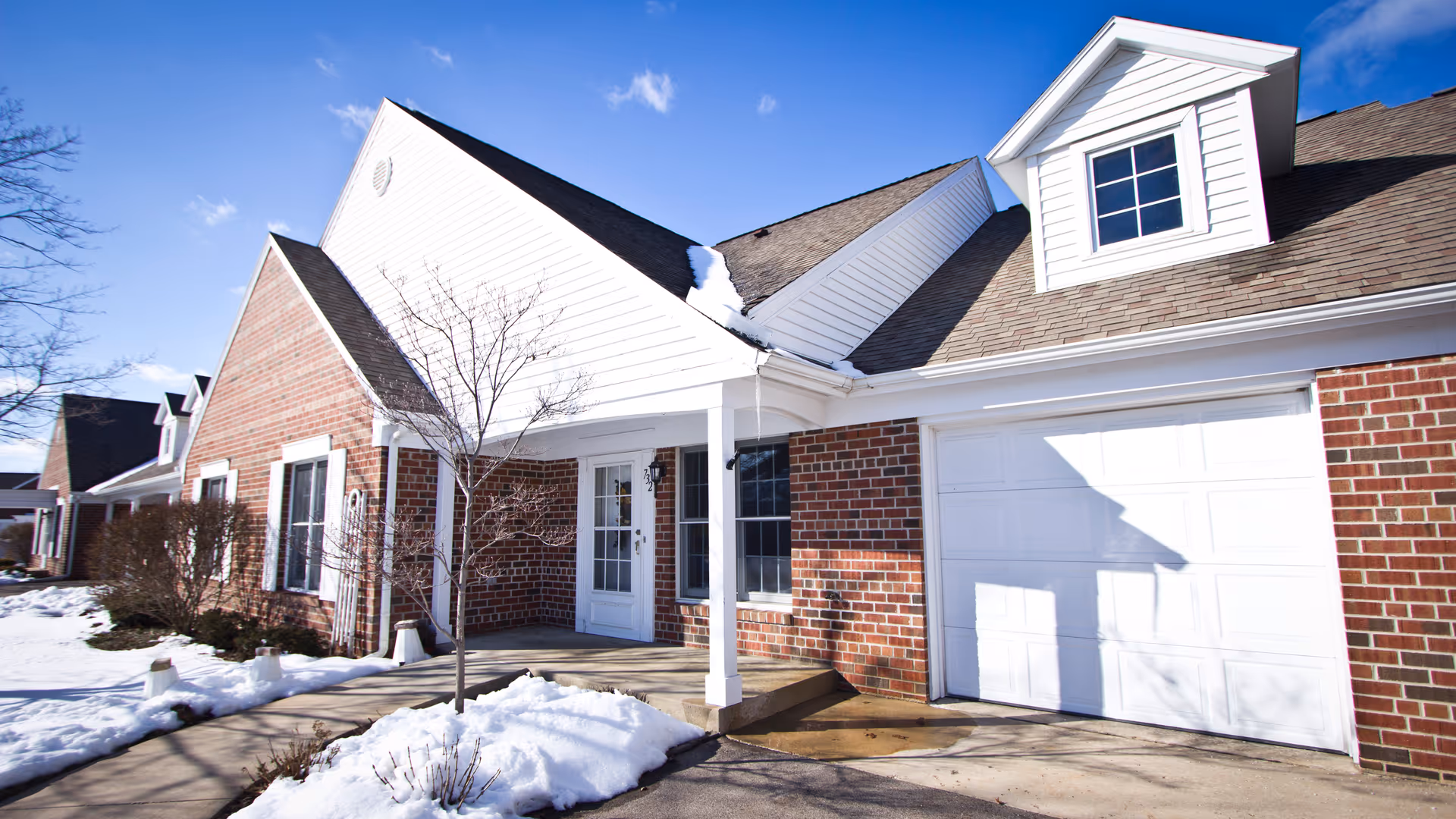 Exterior view of a brick and white siding building with a covered entrance, a garage door, and a small tree in front. Snow is on the ground and the sky is clear and blue.