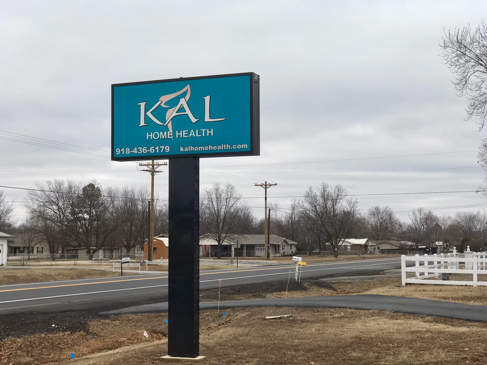 A turquoise KAL Home Health sign on a tall black pole beside a road.