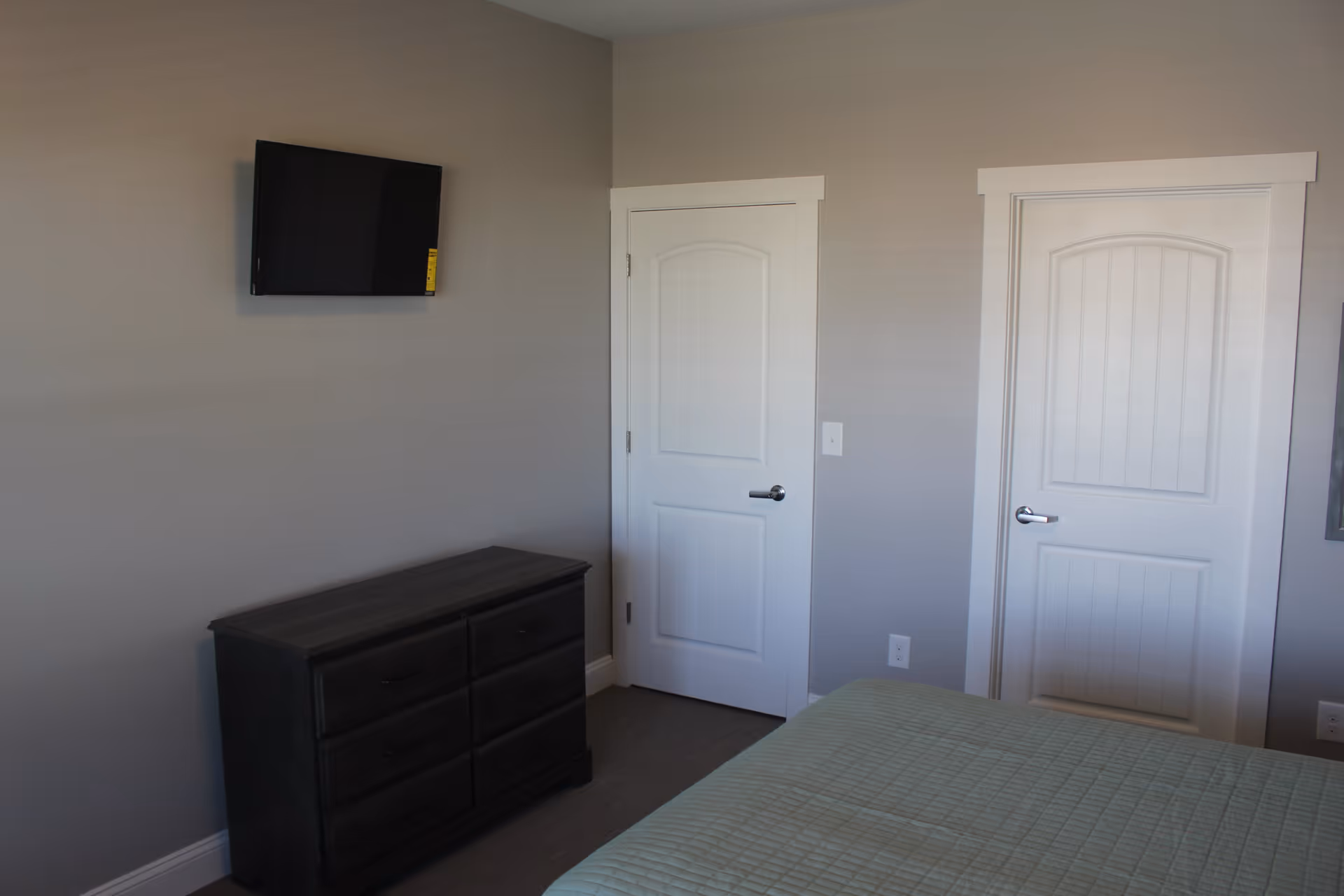 Bedroom with a wall-mounted TV above a dark dresser, two white doors, and part of a bed with a green bedspread.