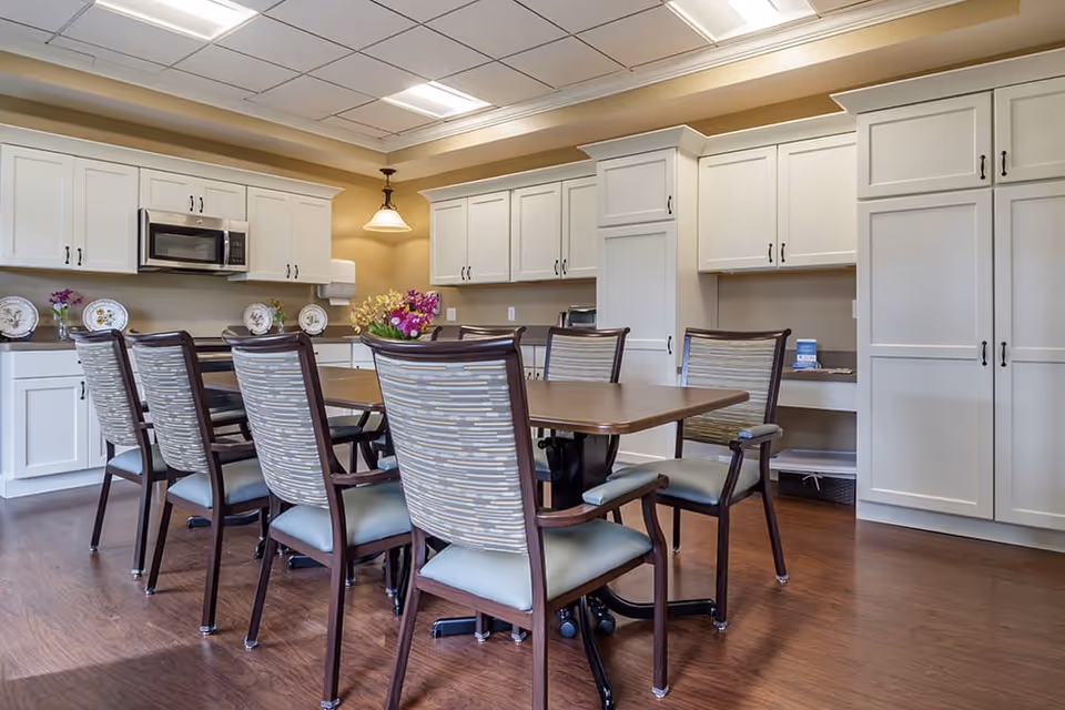A well-lit kitchen area with white cabinets and a wooden table surrounded by eight chairs with patterned backs and cushioned seats. The kitchen features a microwave, countertop with decorative plates and flowers, and a hanging light fixture above the table. The floor is wooden, and the overall setting is clean and organized.
