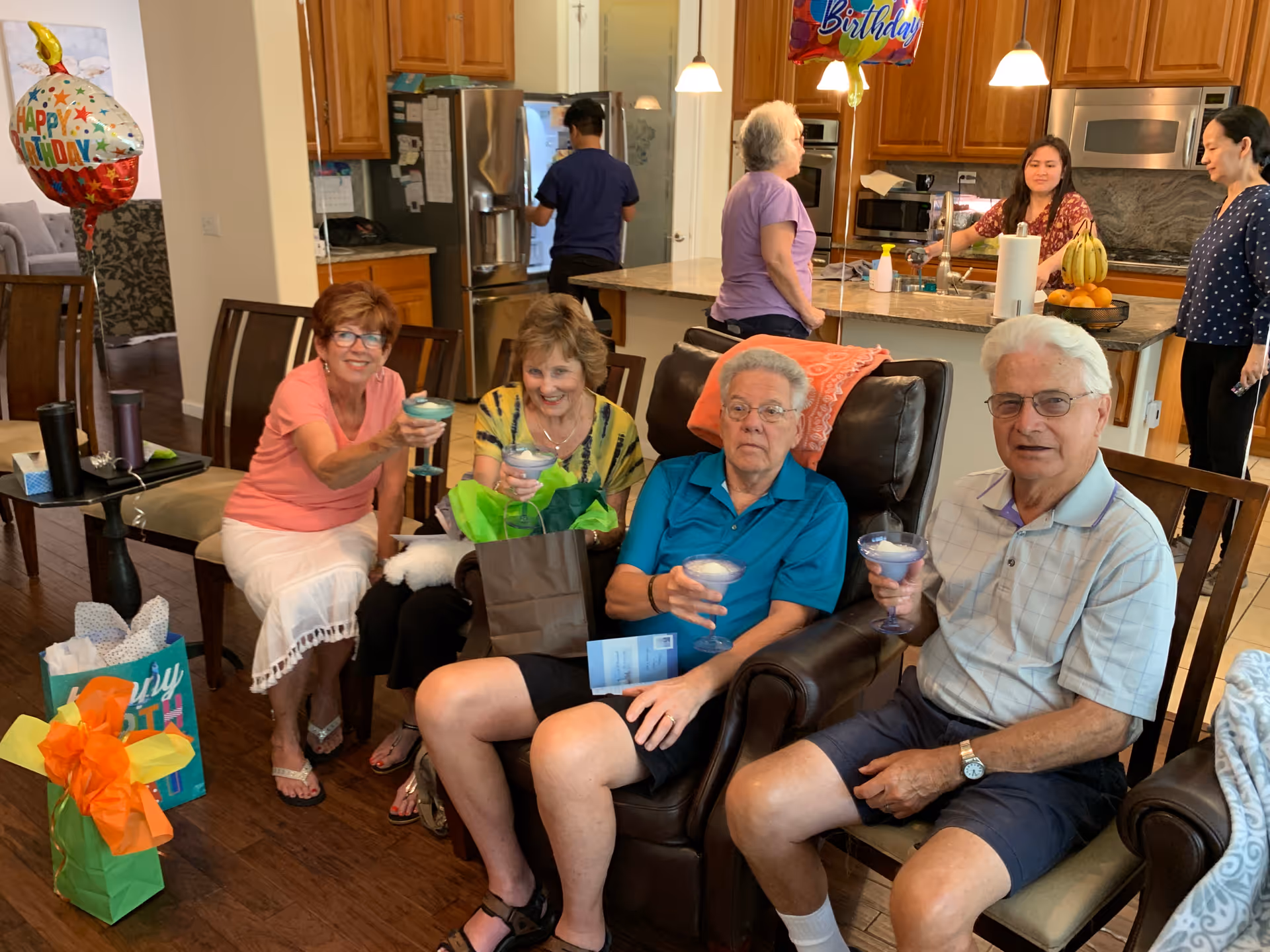 A group of four elderly people sitting in a living room area holding drinks and smiling, with birthday balloons and gift bags nearby. In the background, three adults are standing and talking in a kitchen area with wooden cabinets and stainless steel appliances.
