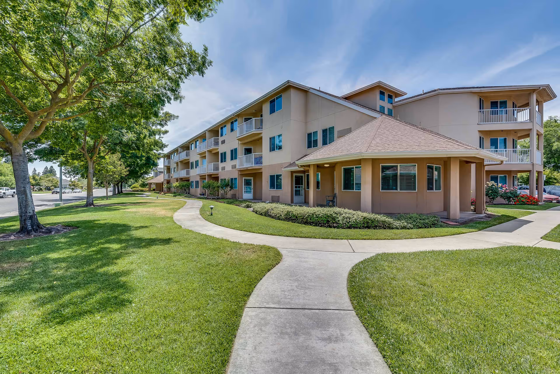 Three-story senior living building with balconies, a covered entrance, curved sidewalk and well-kept lawn under trees.