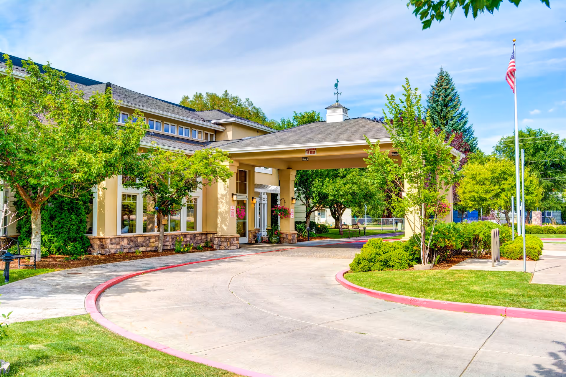 Exterior view of Pelican Pointe by Cogir senior living facility entrance with a covered driveway, surrounded by green trees and landscaping under a partly cloudy sky.