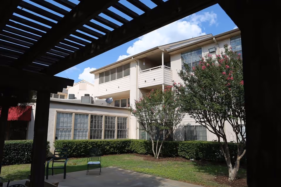 Courtyard view of a multi-story senior living building with balconies, trees, patio chairs and a pergola overhead.