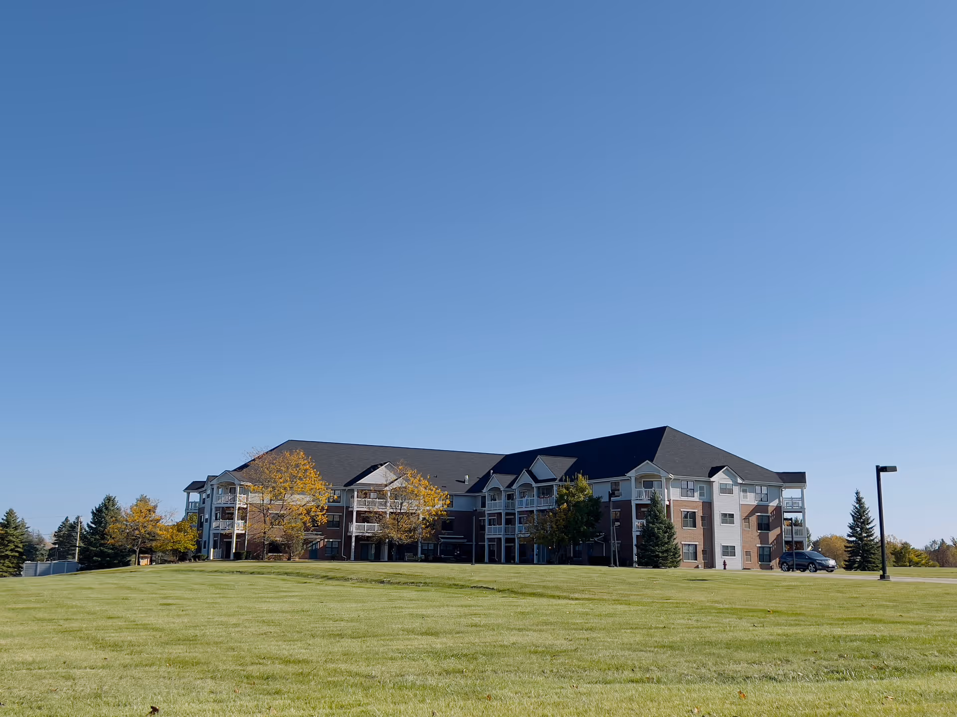 Three-story apartment building with balconies set behind a large grassy lawn under a clear blue sky.
