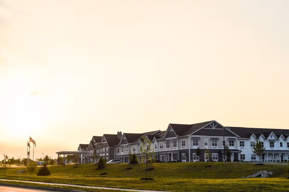Row of connected senior living buildings with manicured lawn and flags at sunset.