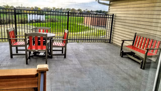 Covered patio with a round table, four red-and-gray chairs and a matching bench behind a metal railing overlooking grass and a pond.