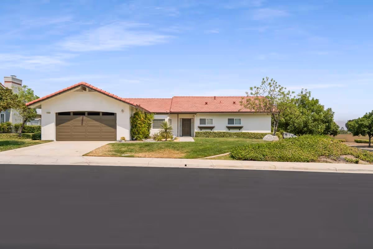 Single-story house with a red tile roof, attached two-car garage, driveway and landscaped front yard under a blue sky.