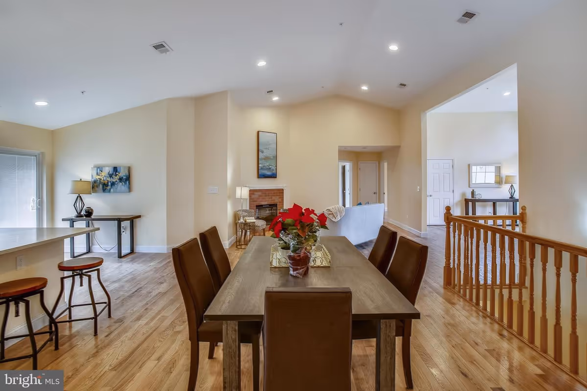 Open-plan dining area with a wooden table and brown chairs overlooking a living room with fireplace, hardwood floors, and a kitchen counter with stools.