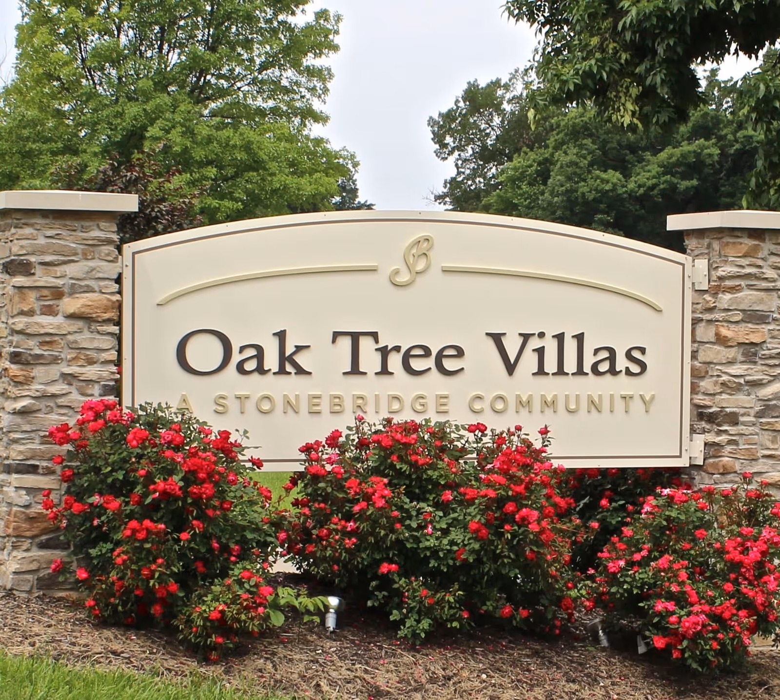 Entrance sign reading "Oak Tree Villas A Stonebridge Community" flanked by stone pillars and red flowering bushes.