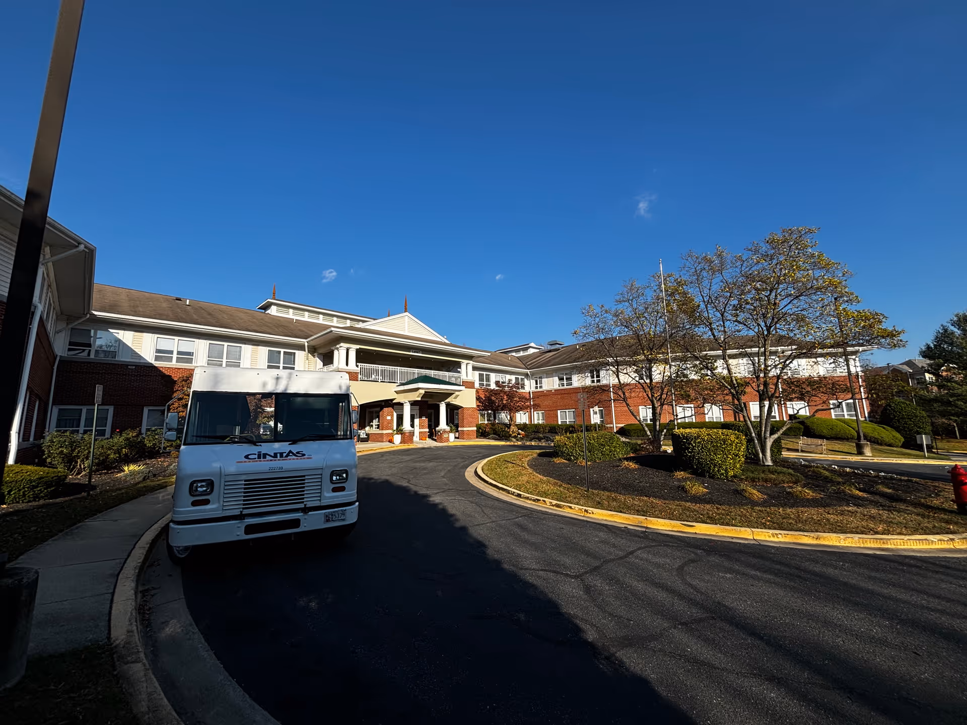 Front exterior of the Brookdale Woodward Estates building with a circular driveway, a delivery truck parked near the entrance, and landscaped grounds under a clear blue sky.