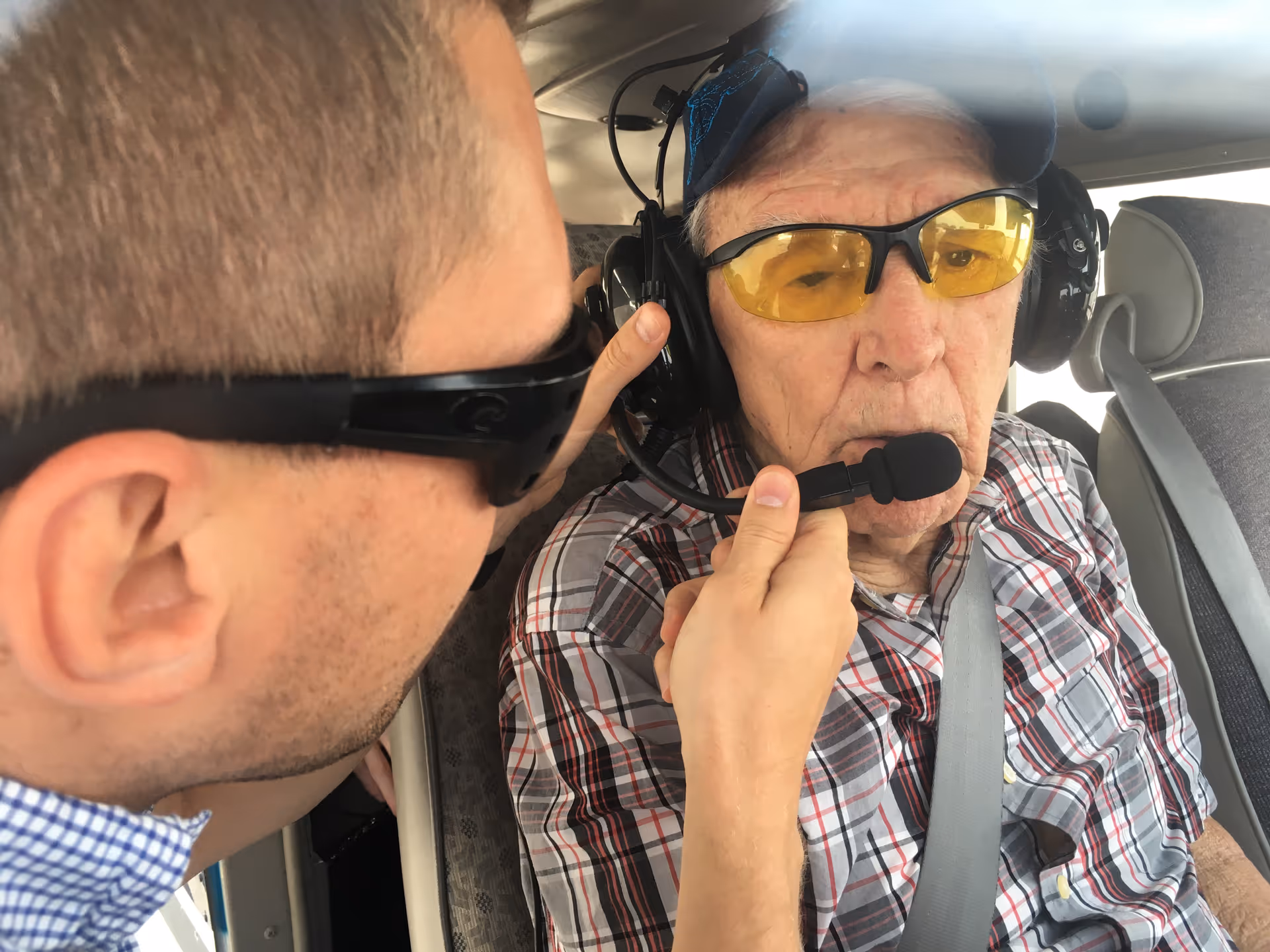 elderly man wearing headset and yellow-tinted glasses, assisted by a younger person inside a small aircraft.
