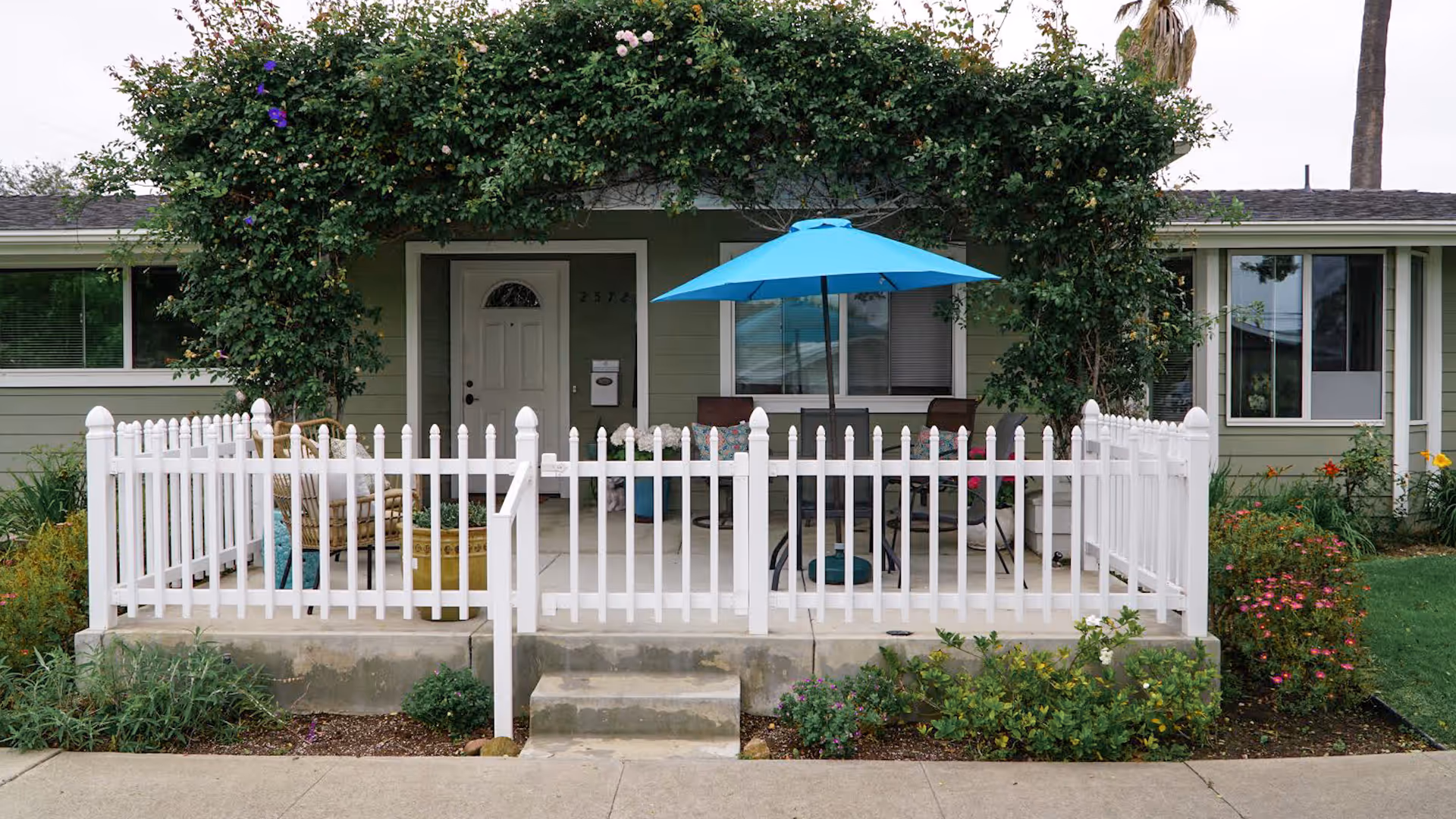 Front porch of a single-story home with a white picket fence, a blue patio umbrella, outdoor chairs, and lush greenery including flowering plants and vines around the entrance.