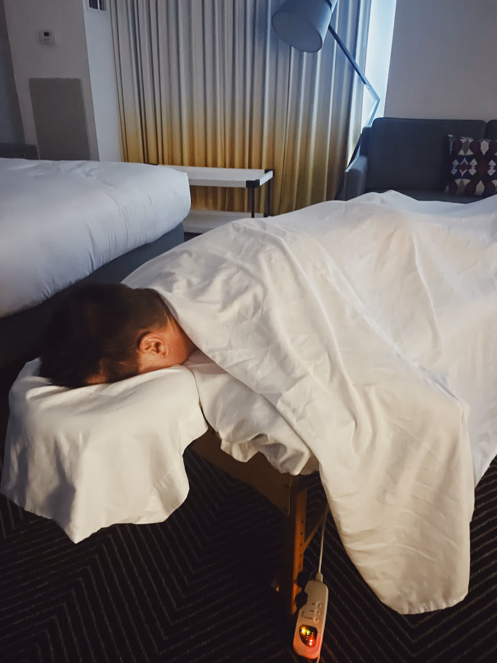 Person resting face down on a white-blanket massage table in a cozy bedroom.