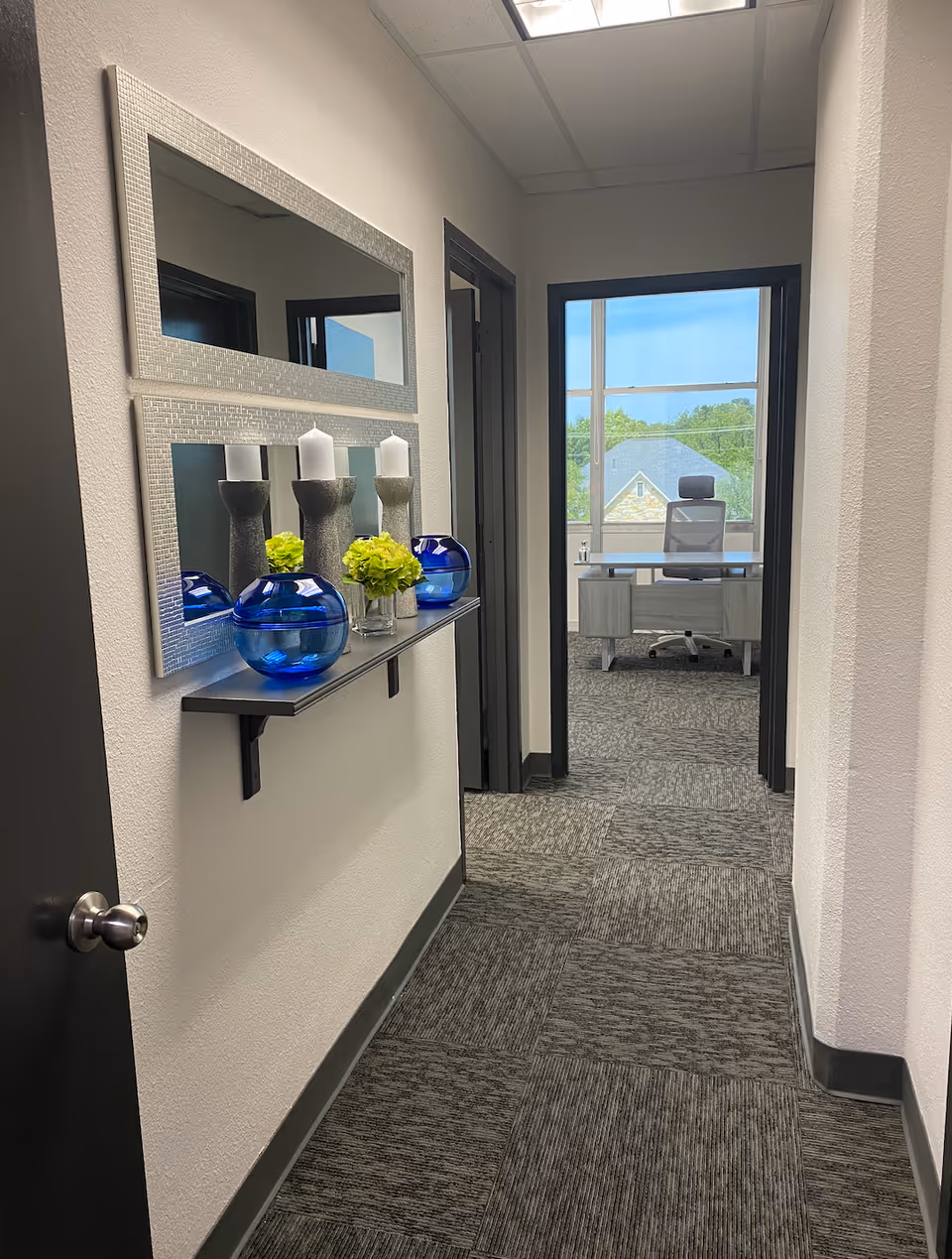 Narrow office hallway with decorative shelf, mirrors, blue vases, leading to a bright desk area.