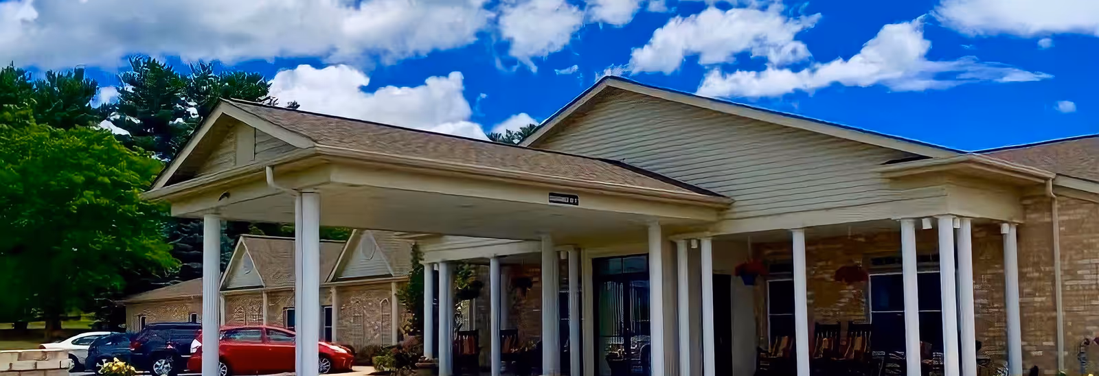 Front entrance of a single-story brick senior living building with a covered porte-cochere supported by white columns and parked cars nearby.