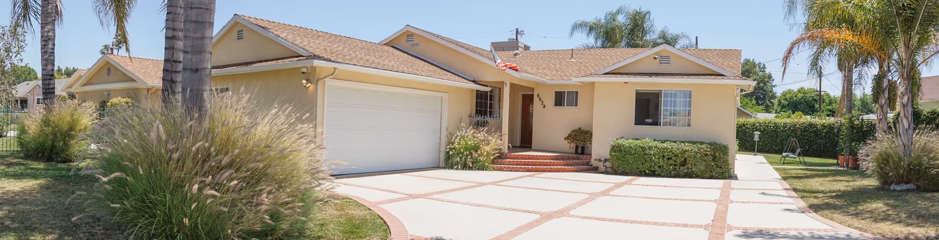 Single-story beige house with a two-car garage, a driveway with a brick pattern, palm trees, and bushes in the front yard under a clear blue sky.