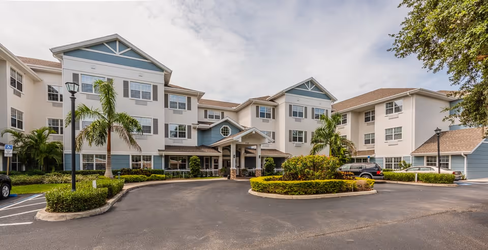 Exterior view of a three-story senior living facility building with a covered entrance, palm trees, and landscaped bushes. The building has white walls with blue accents and multiple windows. There is a paved driveway and parking area in front of the building.
