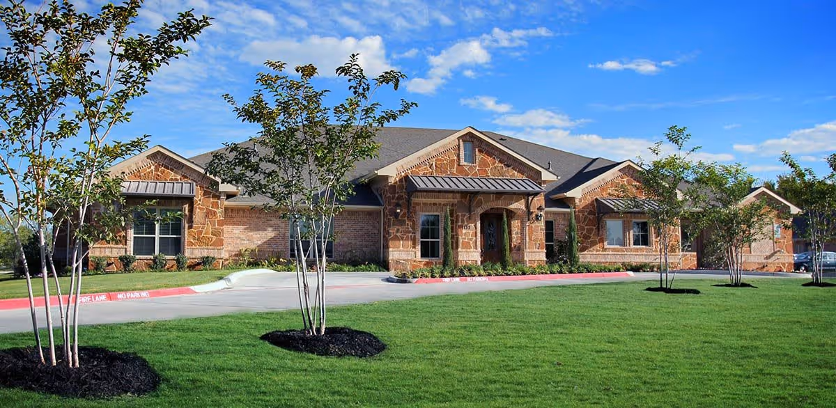Single-story building with stone and brick exterior, surrounded by green lawn and young trees under a blue sky with scattered clouds.