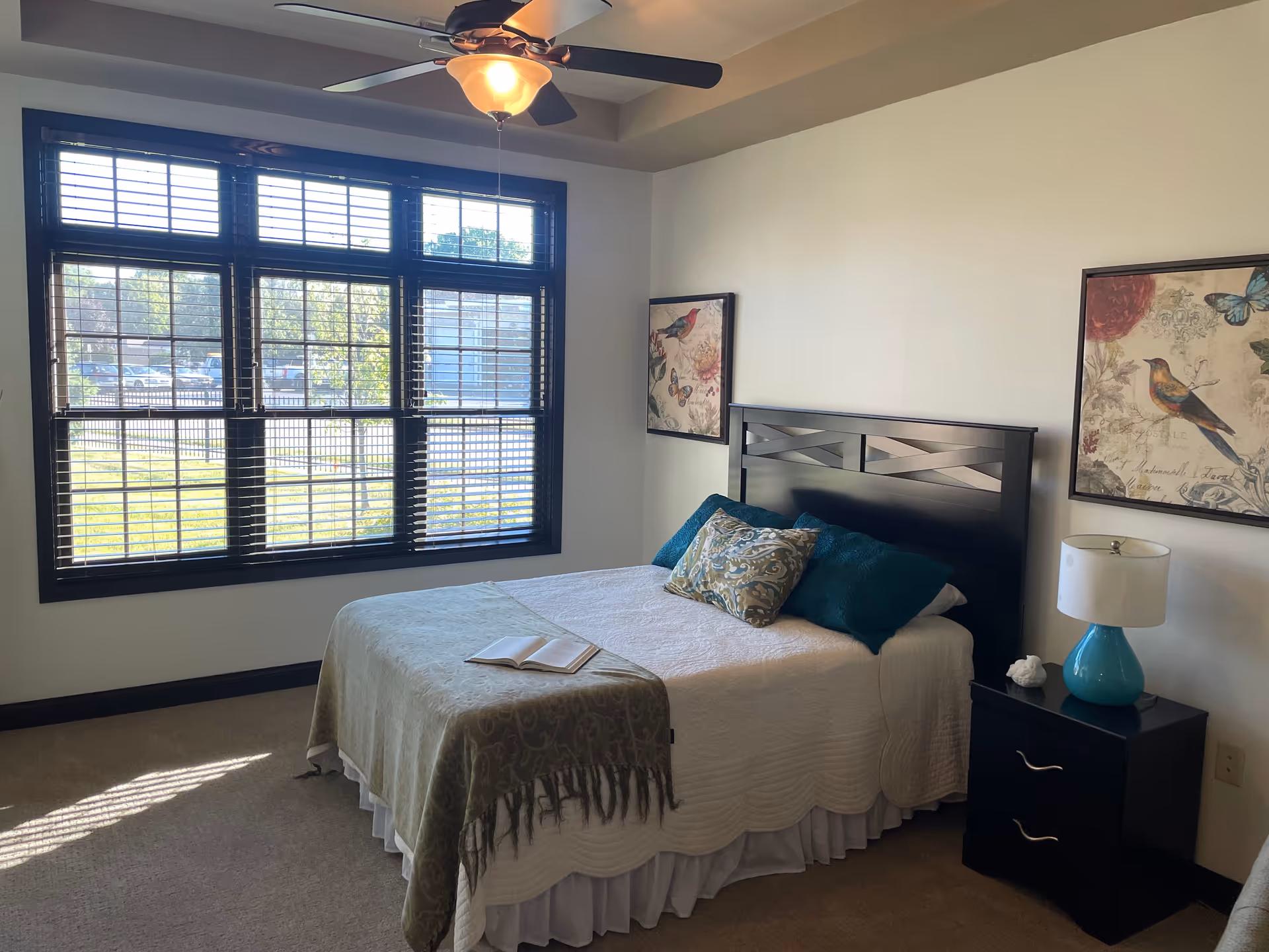 Bright bedroom at Canterbury Arms Assisted Living featuring a large window, bed, nightstand, and teal accents.