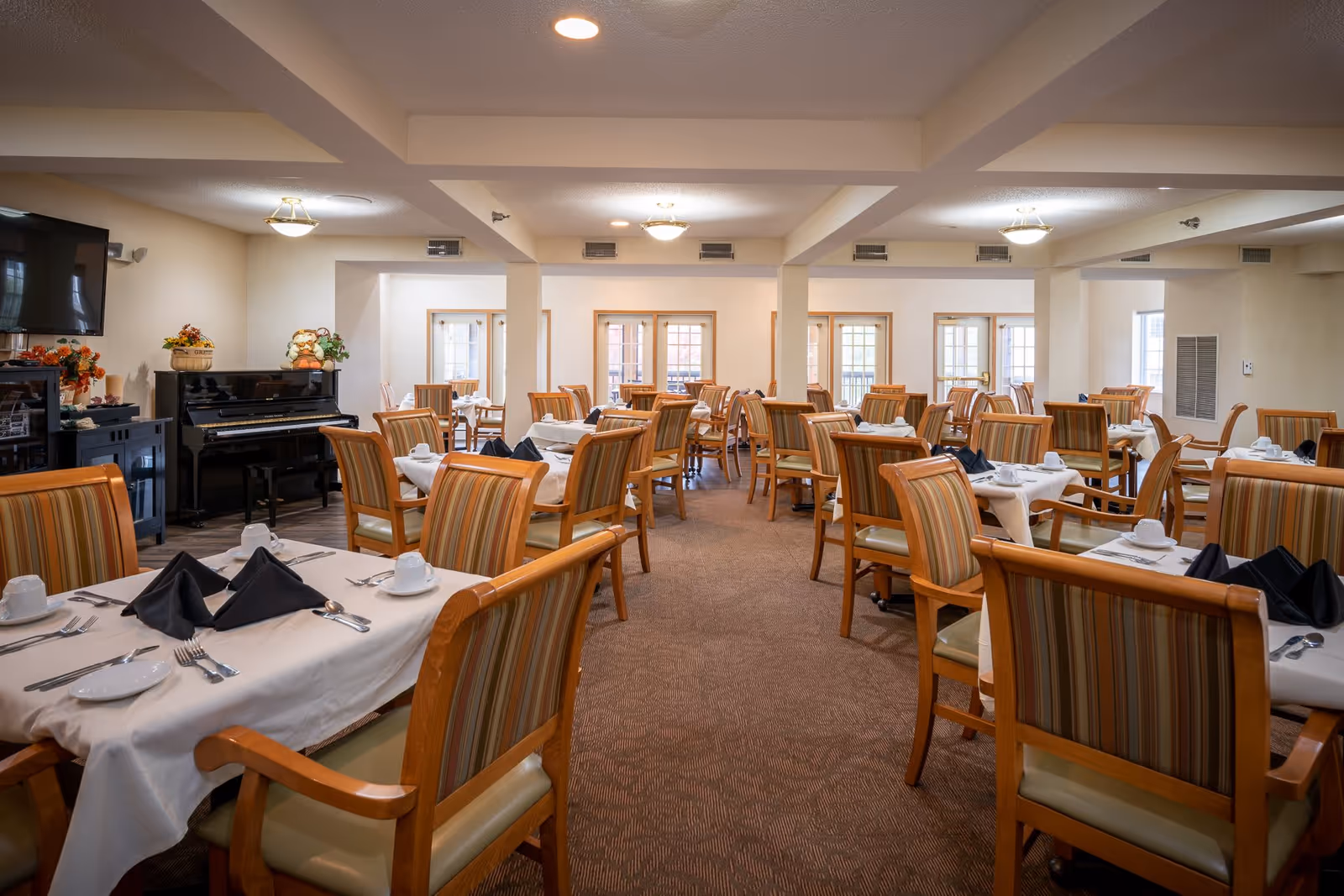 A spacious dining room with multiple tables covered in white tablecloths, each set with plates, cups, silverware, and black napkins. The chairs have wooden frames with striped upholstery. The room is well-lit with ceiling lights and has large windows along the back wall. A piano and some decorative plants are visible on the left side of the room.