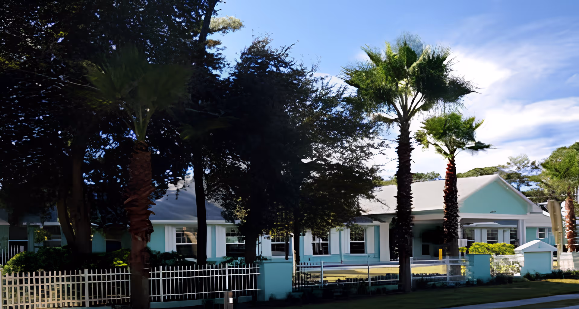 Front exterior of a mint-green assisted living building with palm trees, a white fence, and a clear blue sky.