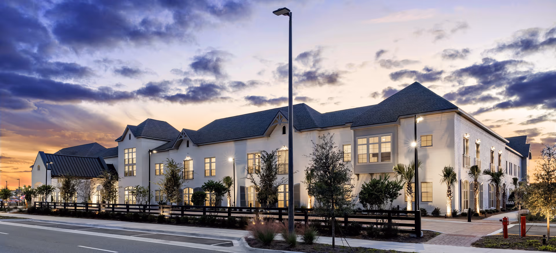 Exterior view of a large, two-story senior living facility building at dusk with lights illuminating the windows and surrounding landscaping. The sky is partly cloudy with a sunset glow. There are trees, shrubs, and a wooden fence along the sidewalk in front of the building.