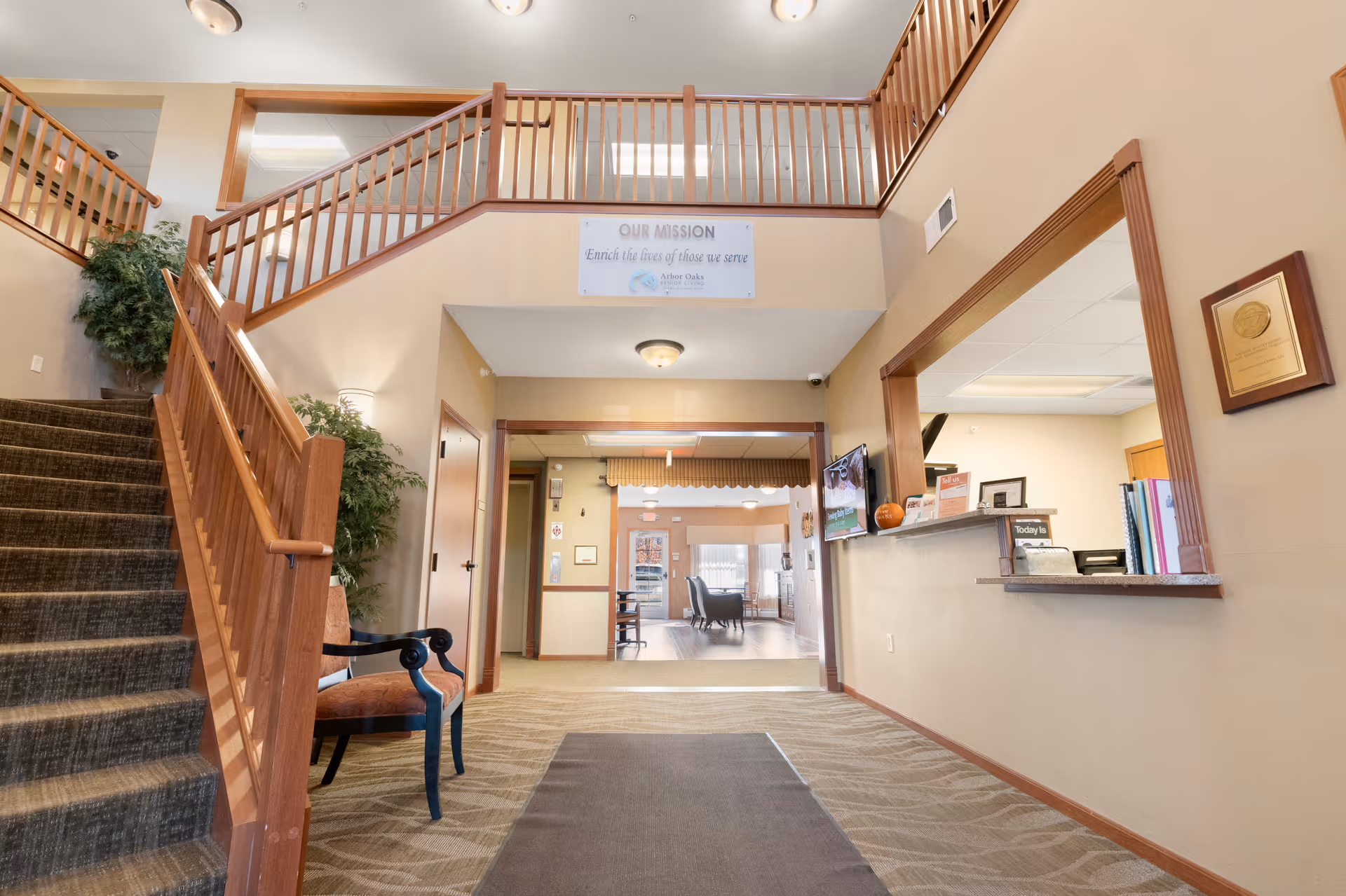 Lobby of a senior living facility showing a carpeted entry with a staircase, reception desk, and seating area.