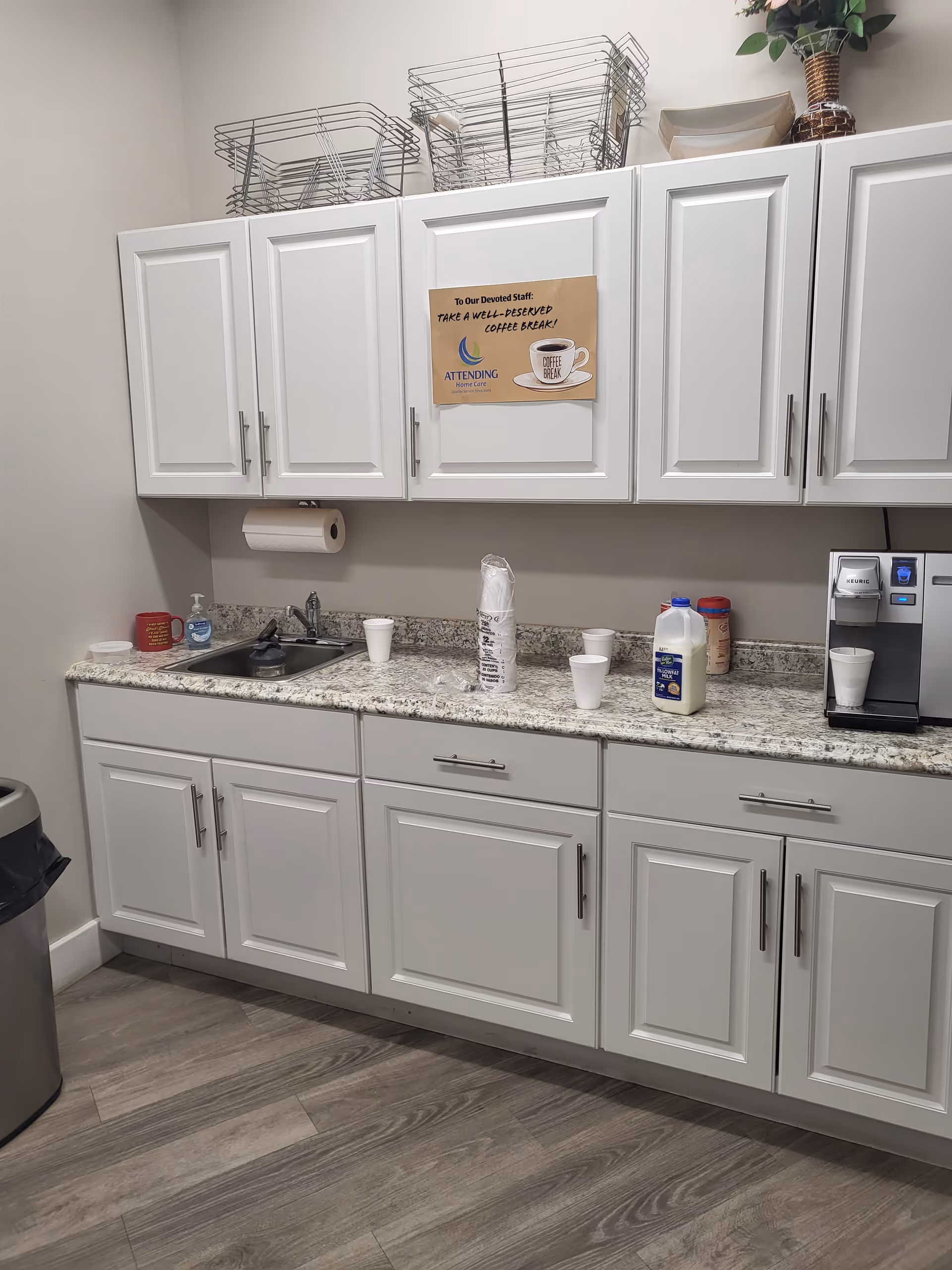 A clean kitchenette with white cabinets, granite counter, coffee station, and dish racks.