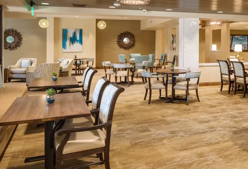 Spacious dining and lounge area with multiple round and rectangular tables, upholstered chairs, armchairs, and decorative wall art under warm lighting.