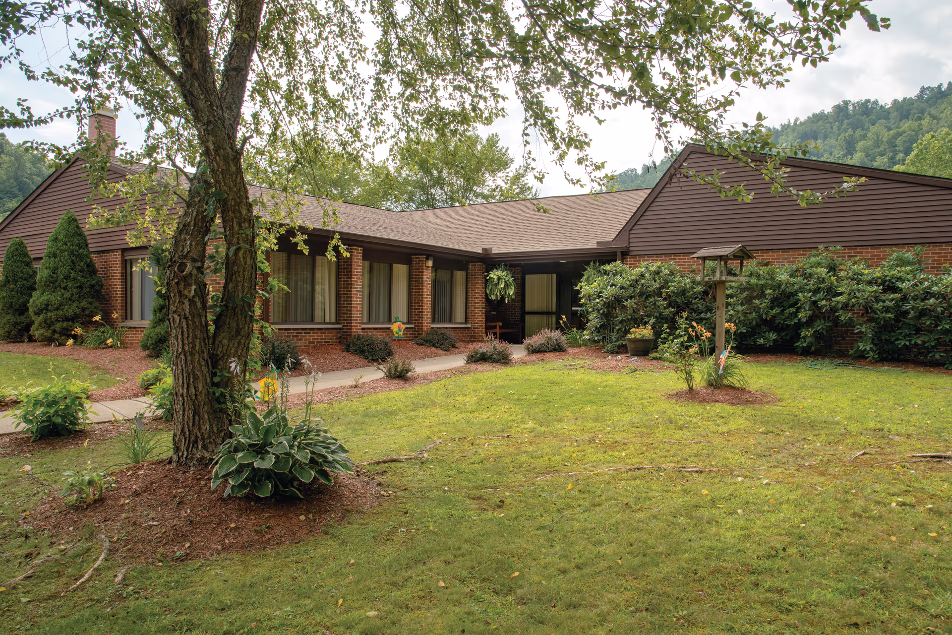 Exterior view of a single-story brick building with brown siding and a shingled roof, surrounded by green grass, shrubs, and trees, with a mountainous background under a cloudy sky.