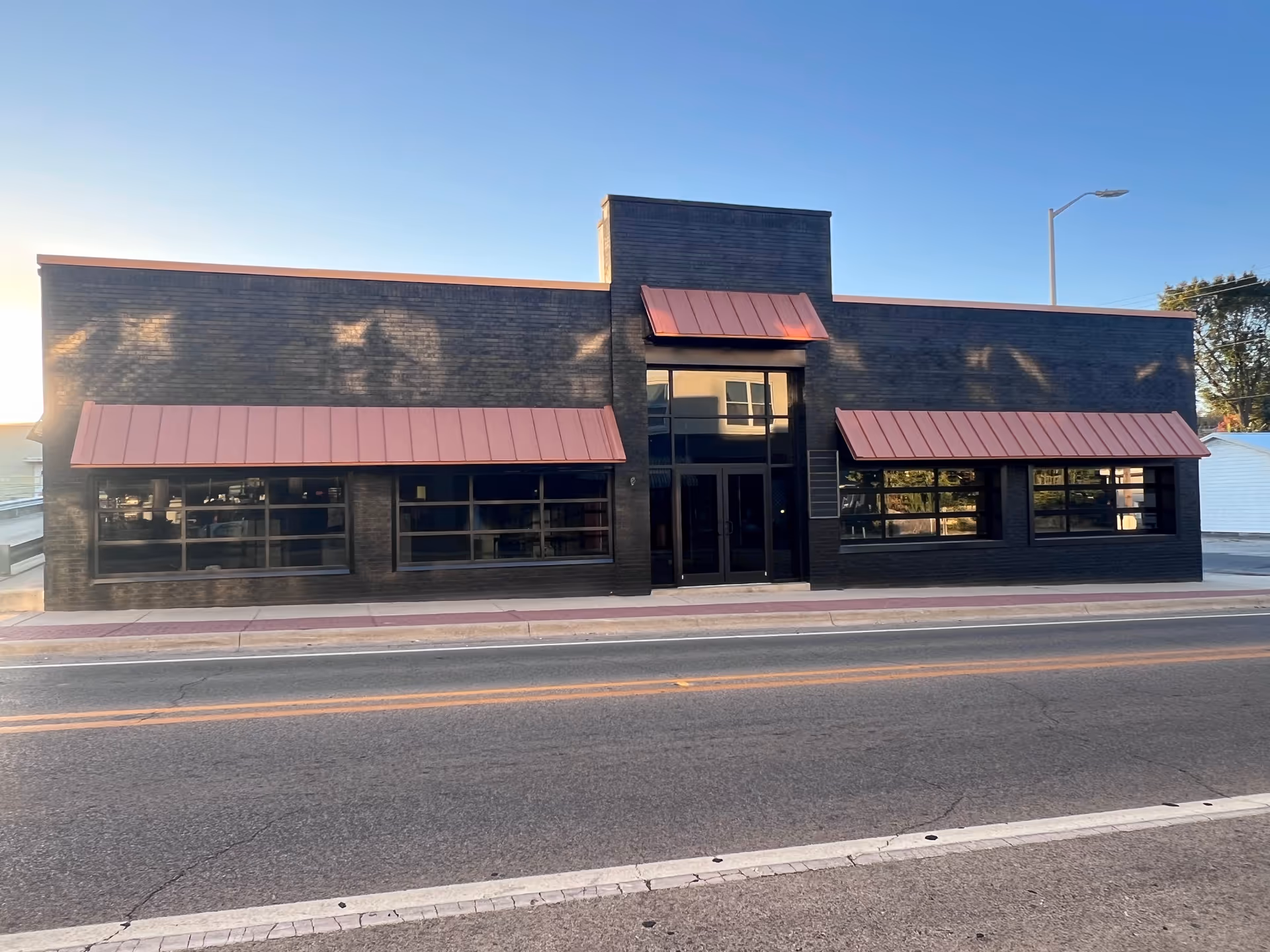 A black brick building with red awnings and large front windows along a street.