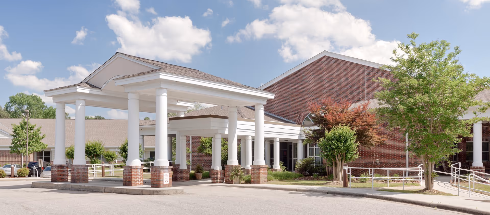 Brick senior living facility exterior with white columned entrance and landscaped driveway.