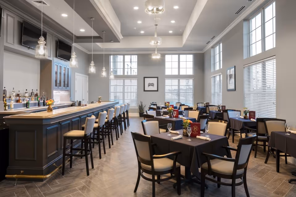A bright and spacious dining room with multiple tables covered in black tablecloths, each set with glasses, napkins, and small flower arrangements. There is a long bar with high chairs on the left side, pendant lights hanging above, and large windows letting in natural light.