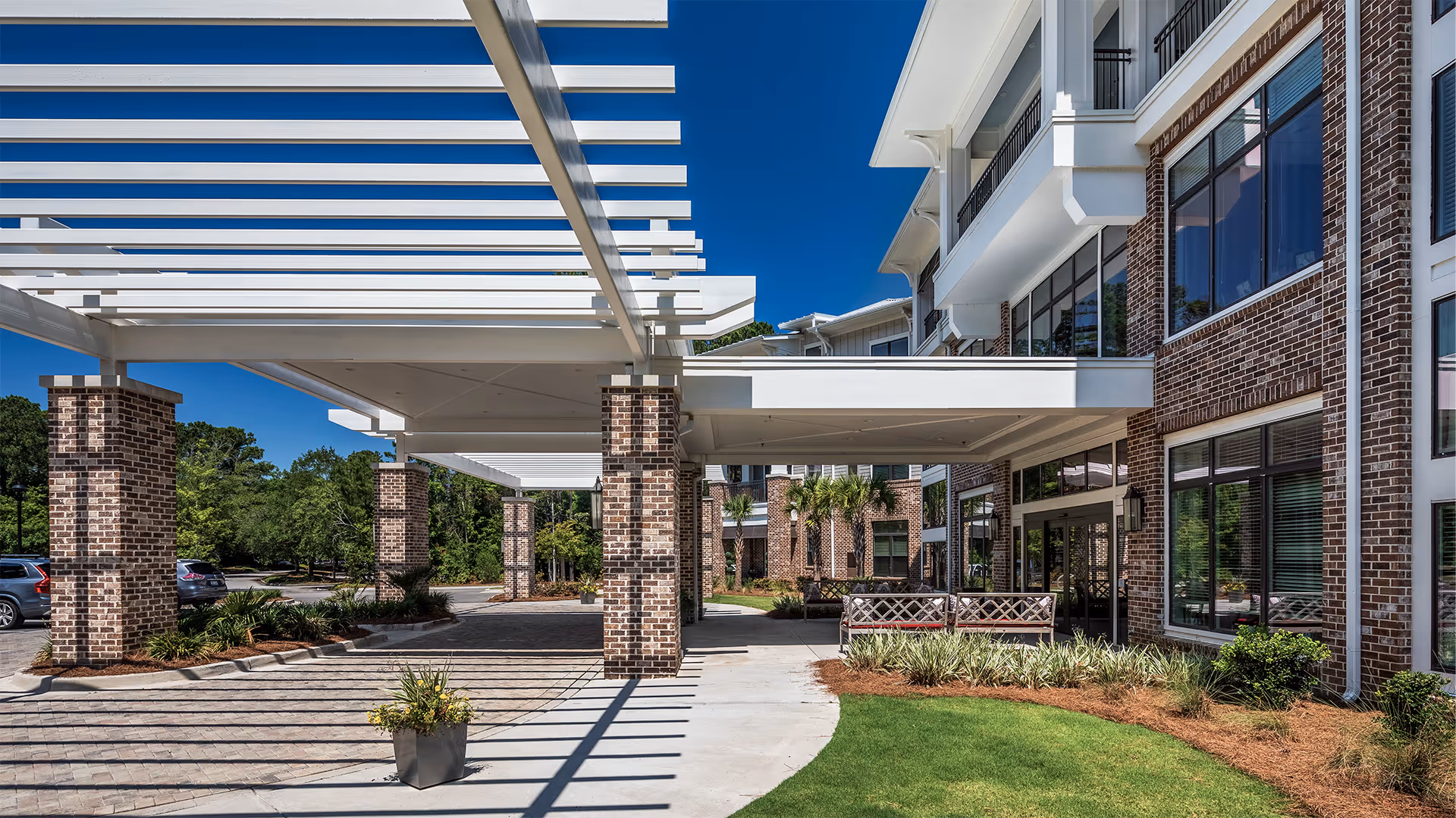 Entrance area of The Artisan Mount Pleasant senior living facility featuring a covered drop-off zone with brick pillars and white pergola-style beams, adjacent to a well-maintained lawn and landscaped garden beds under a clear blue sky.