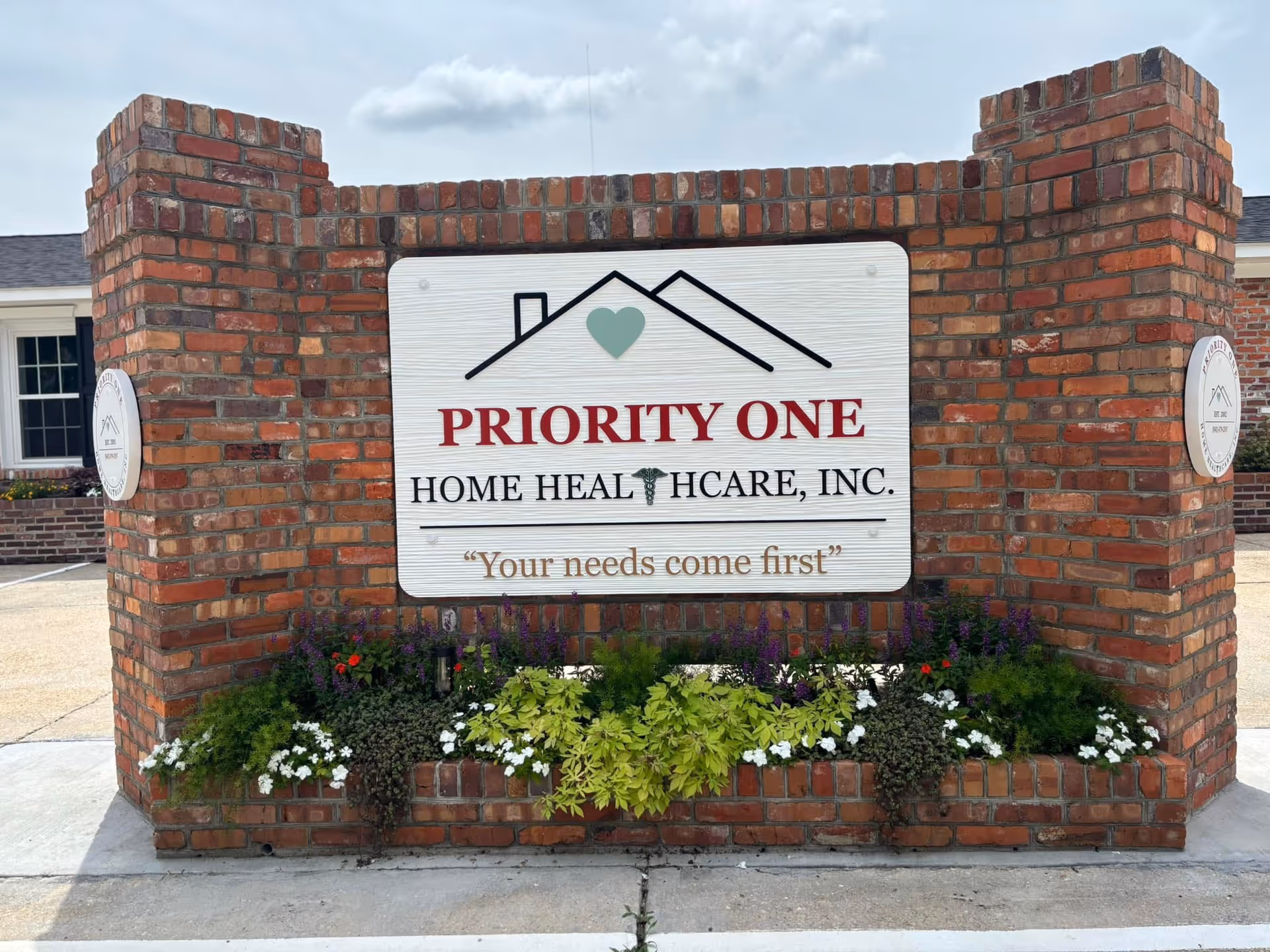 Brick entrance with a sign reading Priority One Home Healthcare, Inc., surrounded by flowers.
