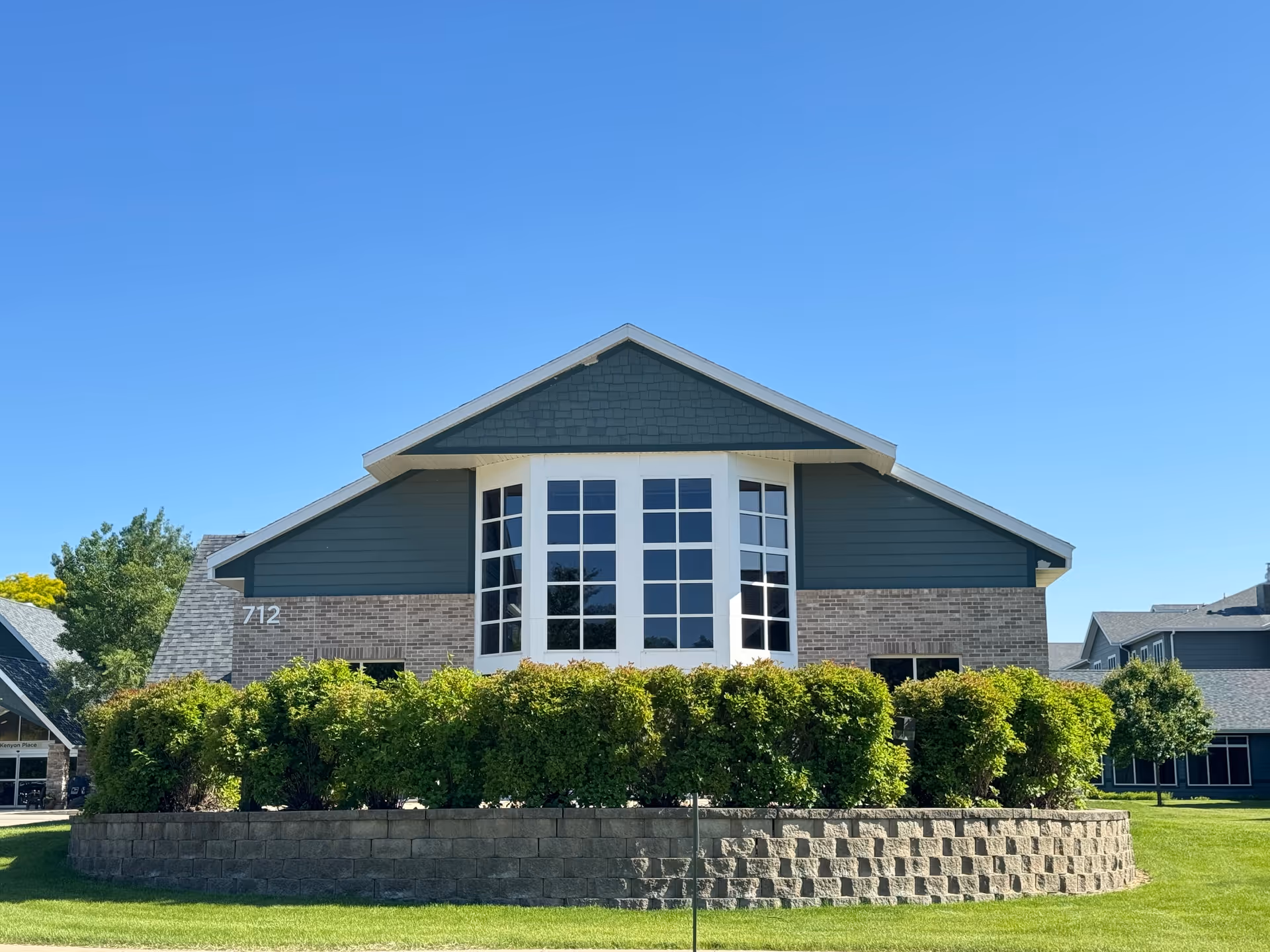 Front exterior view of a senior living facility building with large windows, green siding, brick accents, and a neatly trimmed hedge in front under a clear blue sky.