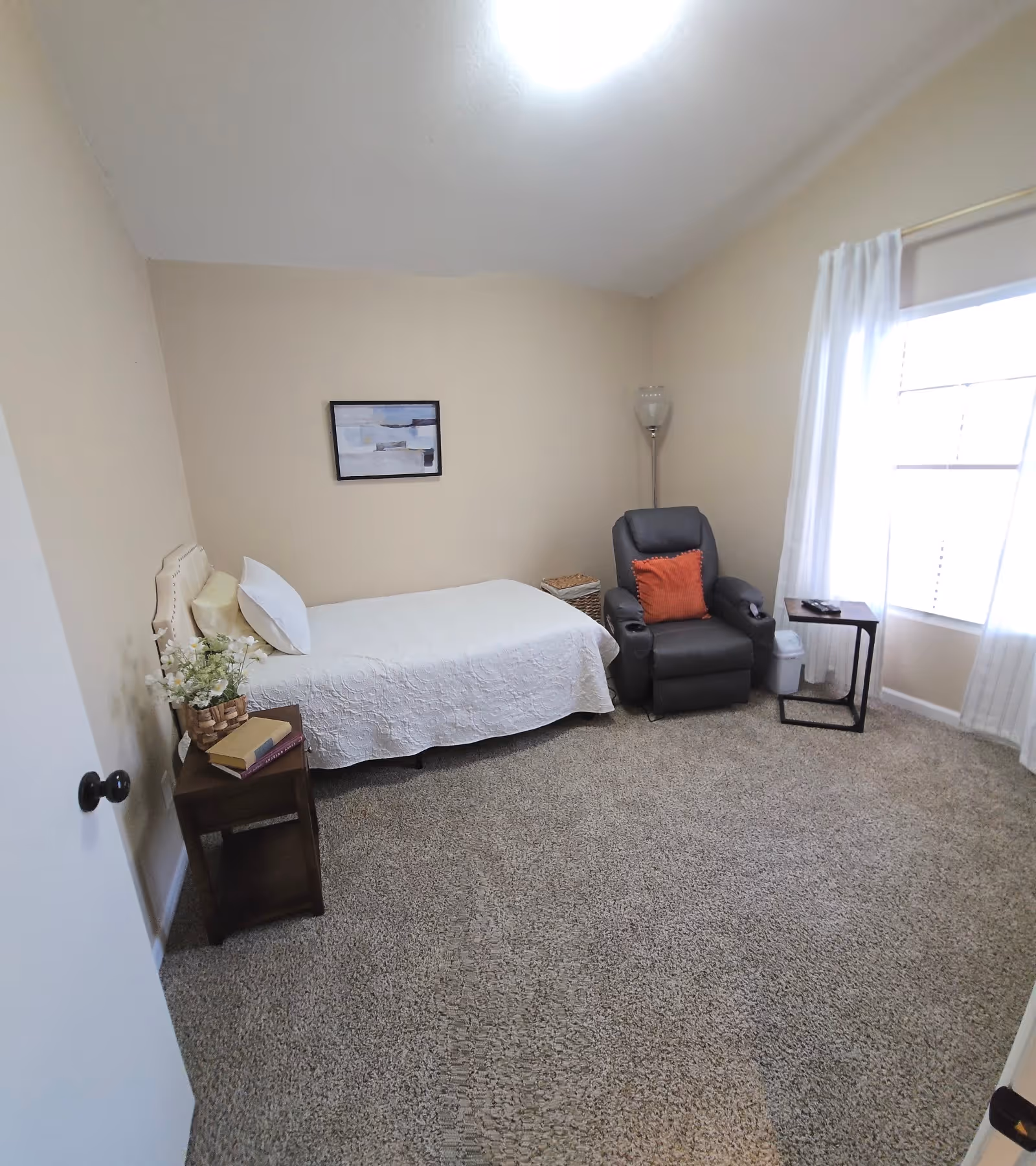 Small bedroom with a single bed, recliner, side table, and window with white curtains.