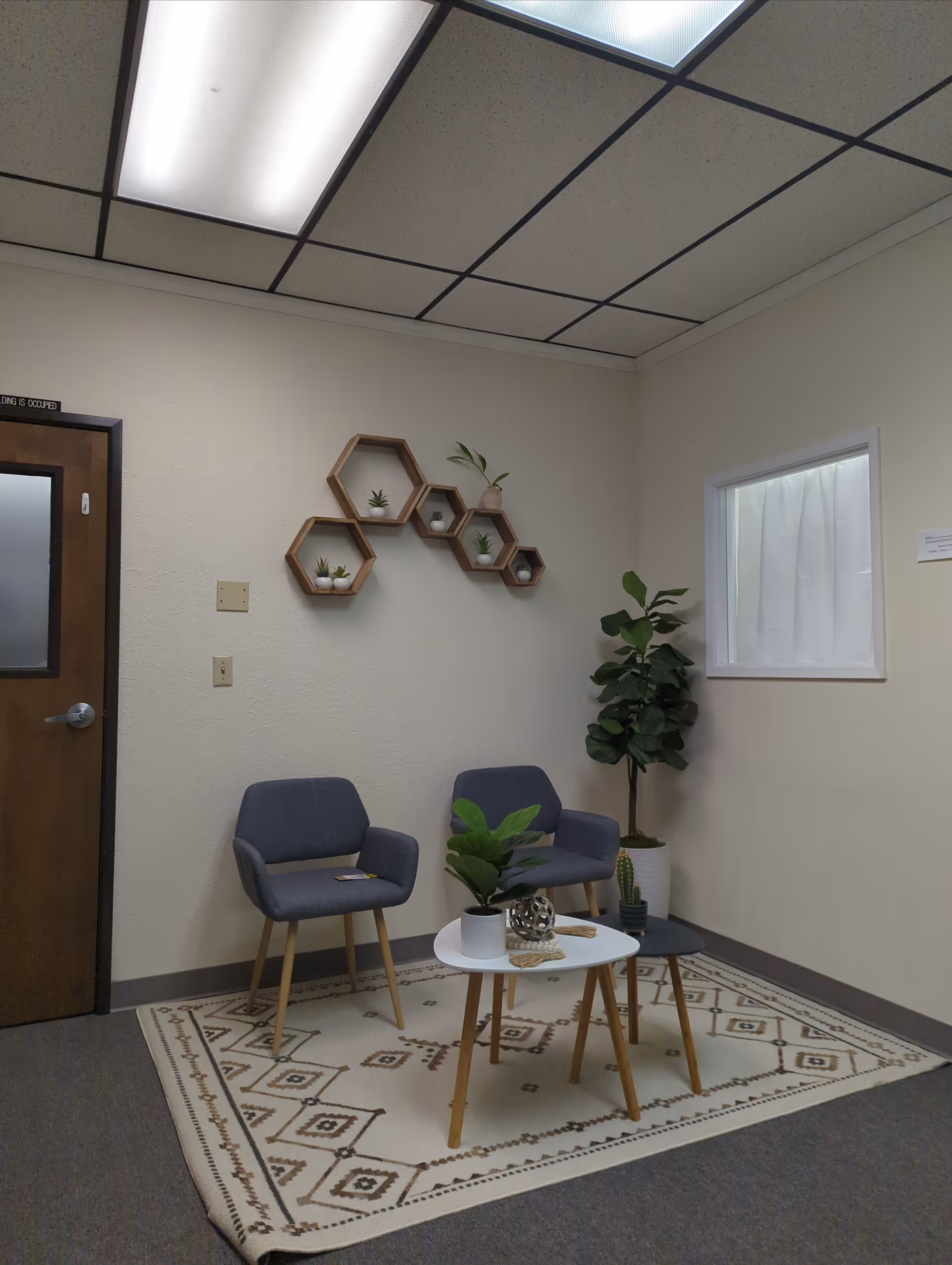 Interior lounge area with two gray chairs, a white round table, shelves, and plants.