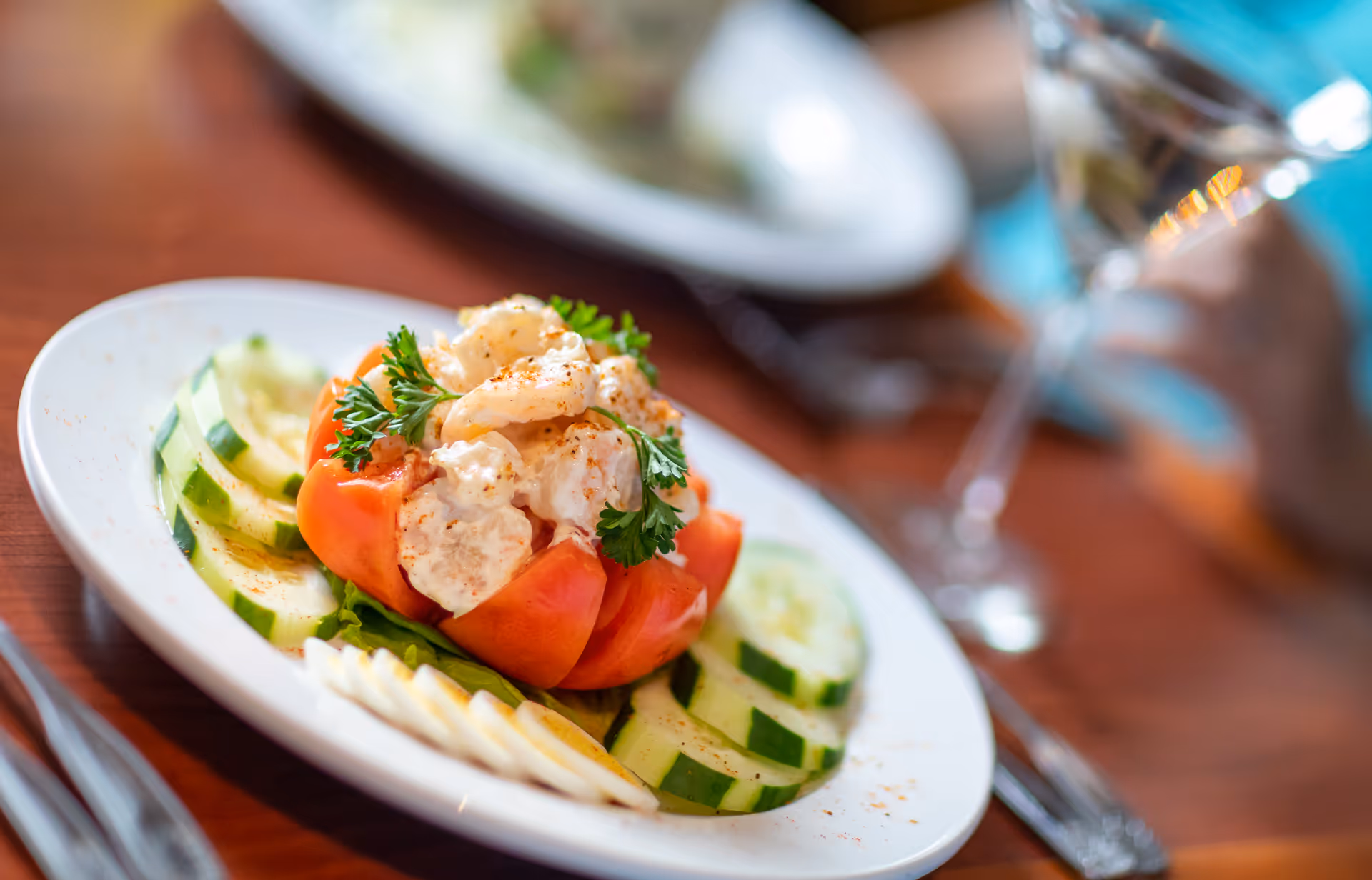 A close-up of a plated shrimp salad served in a tomato bowl, garnished with parsley, surrounded by sliced cucumbers and hard-boiled egg slices on a white plate, with a glass of water and utensils on a wooden table in the background.