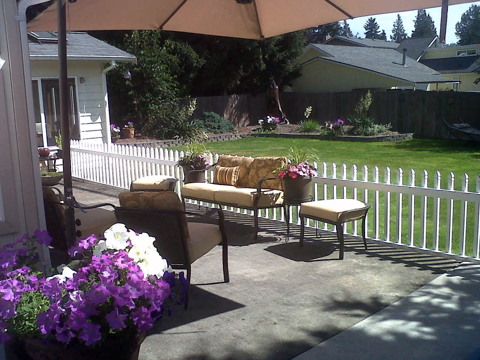 Sunlit backyard patio with cushioned outdoor seating, potted flowers, a white picket fence and a green lawn.