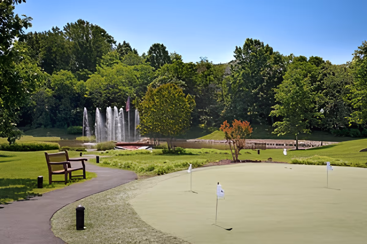 A peaceful outdoor scene at Belvoir Woods Health Care Center featuring a putting green with several golf flags, a paved walking path with a wooden bench, a water fountain spraying multiple streams of water, and lush green trees under a clear blue sky.