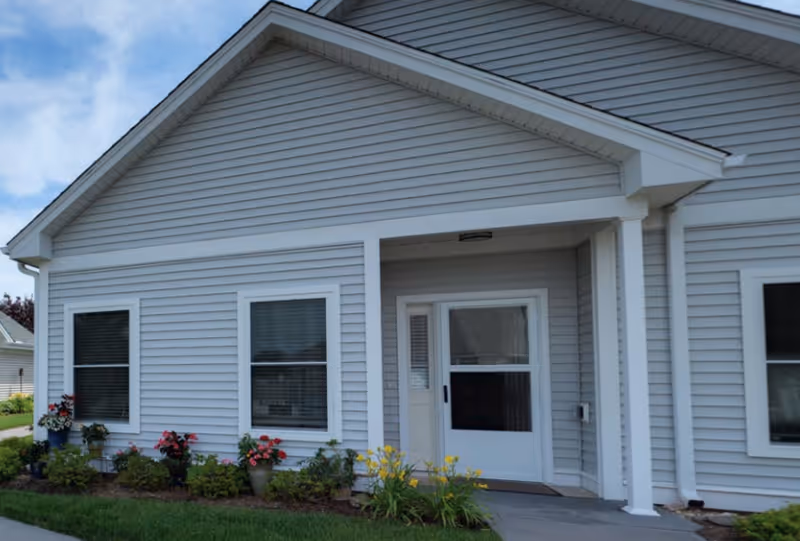Front entrance of a single-story light-gray siding building with a small covered porch, white door and windows and flower beds along the foundation.