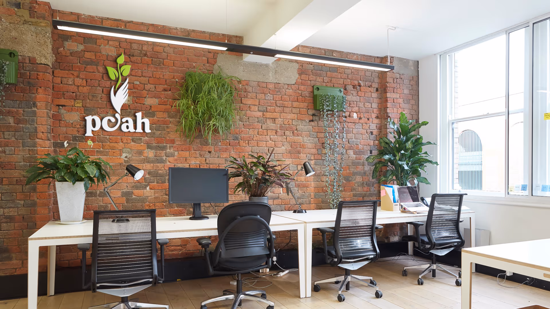 Open-office workspace with a brick wall, desks, chairs, plants, and a logo.