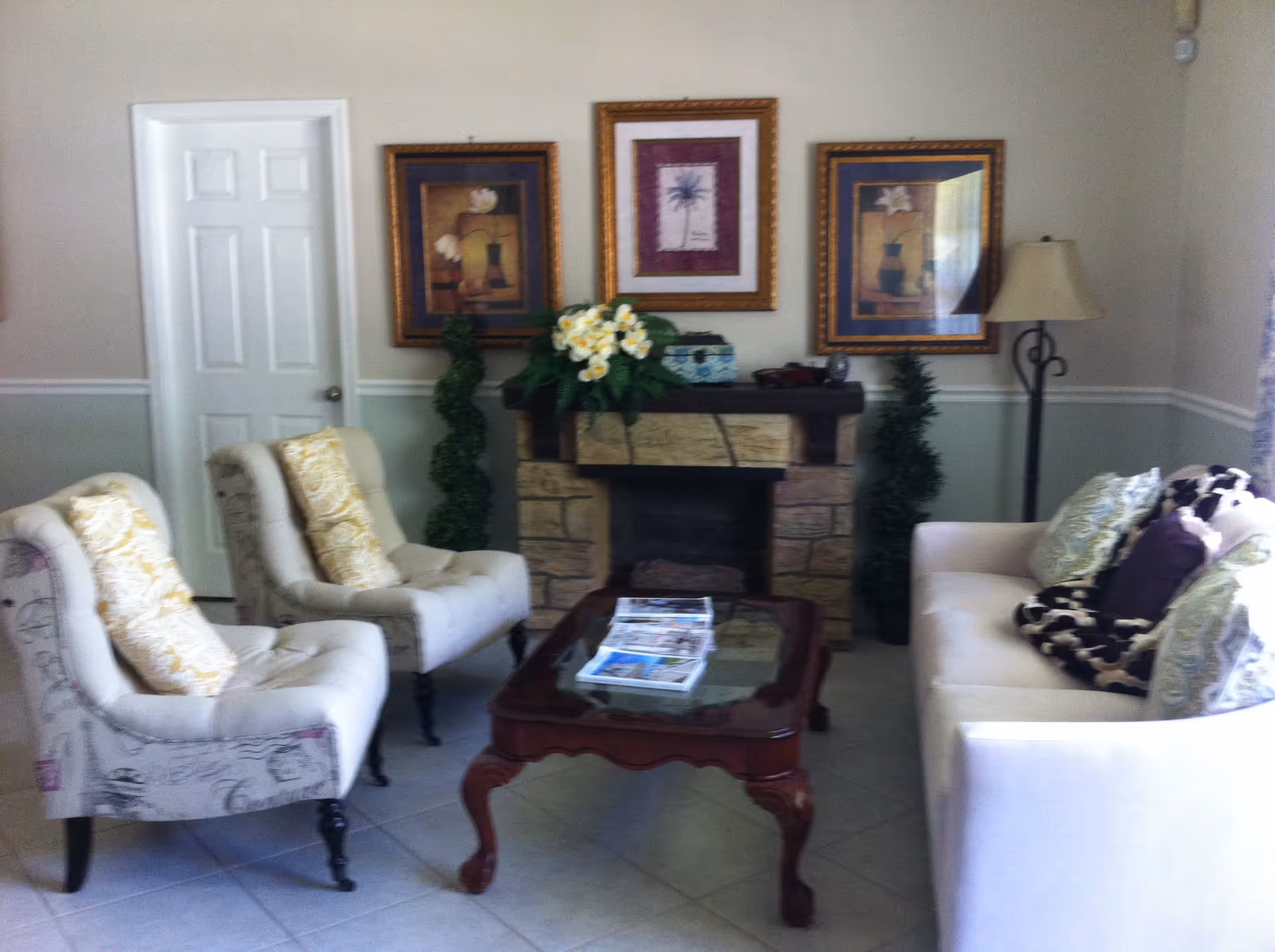 A cozy living room with a white sofa adorned with patterned pillows and a throw blanket, two upholstered armchairs with yellow patterned cushions, a wooden coffee table with magazines on it, a stone fireplace decorated with a flower arrangement and two tall green plants on either side, three framed artworks hanging above the fireplace, and a floor lamp in the corner.