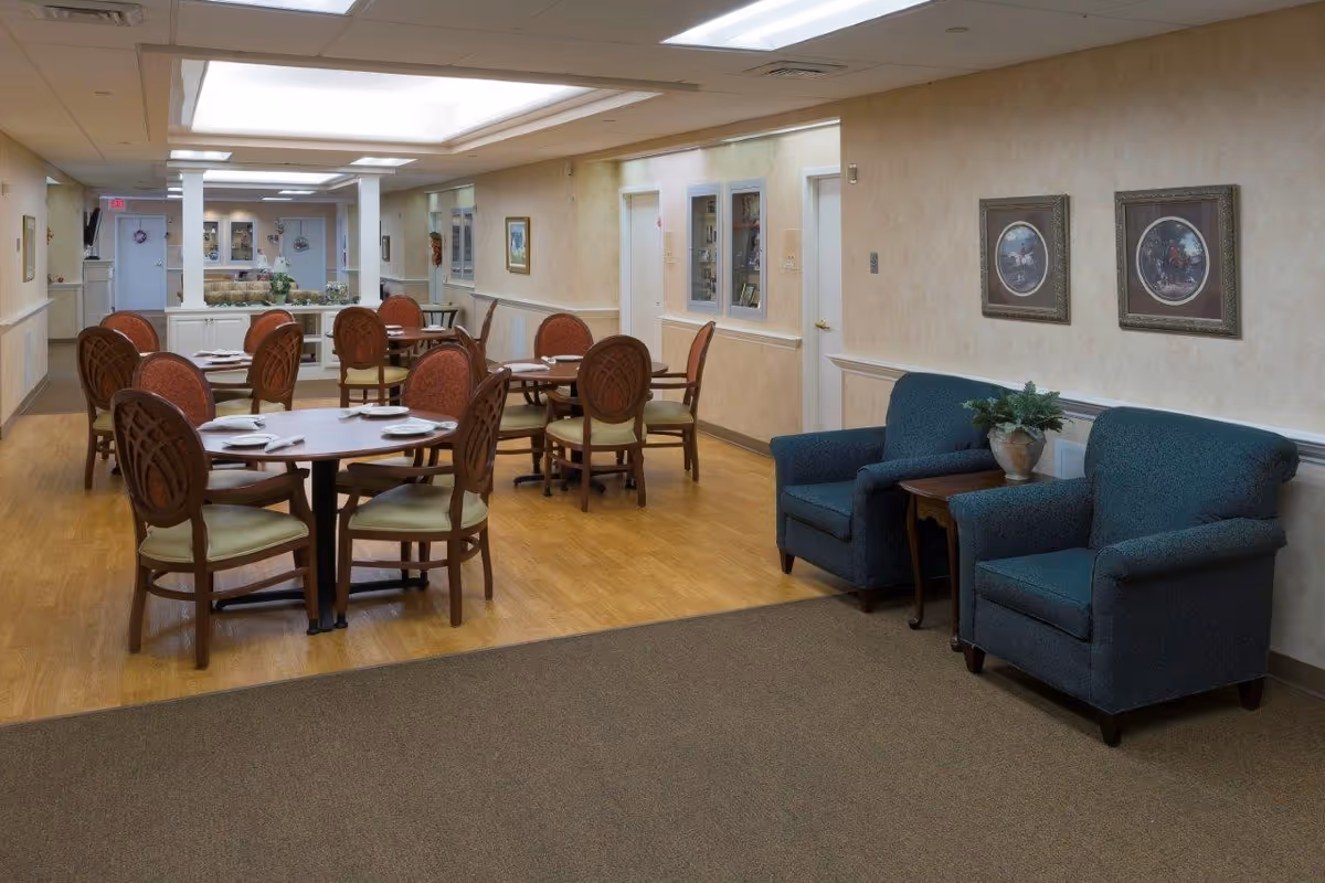 Dining room in a senior living facility with round tables set for meals, multiple chairs, and two blue armchairs by the wall.