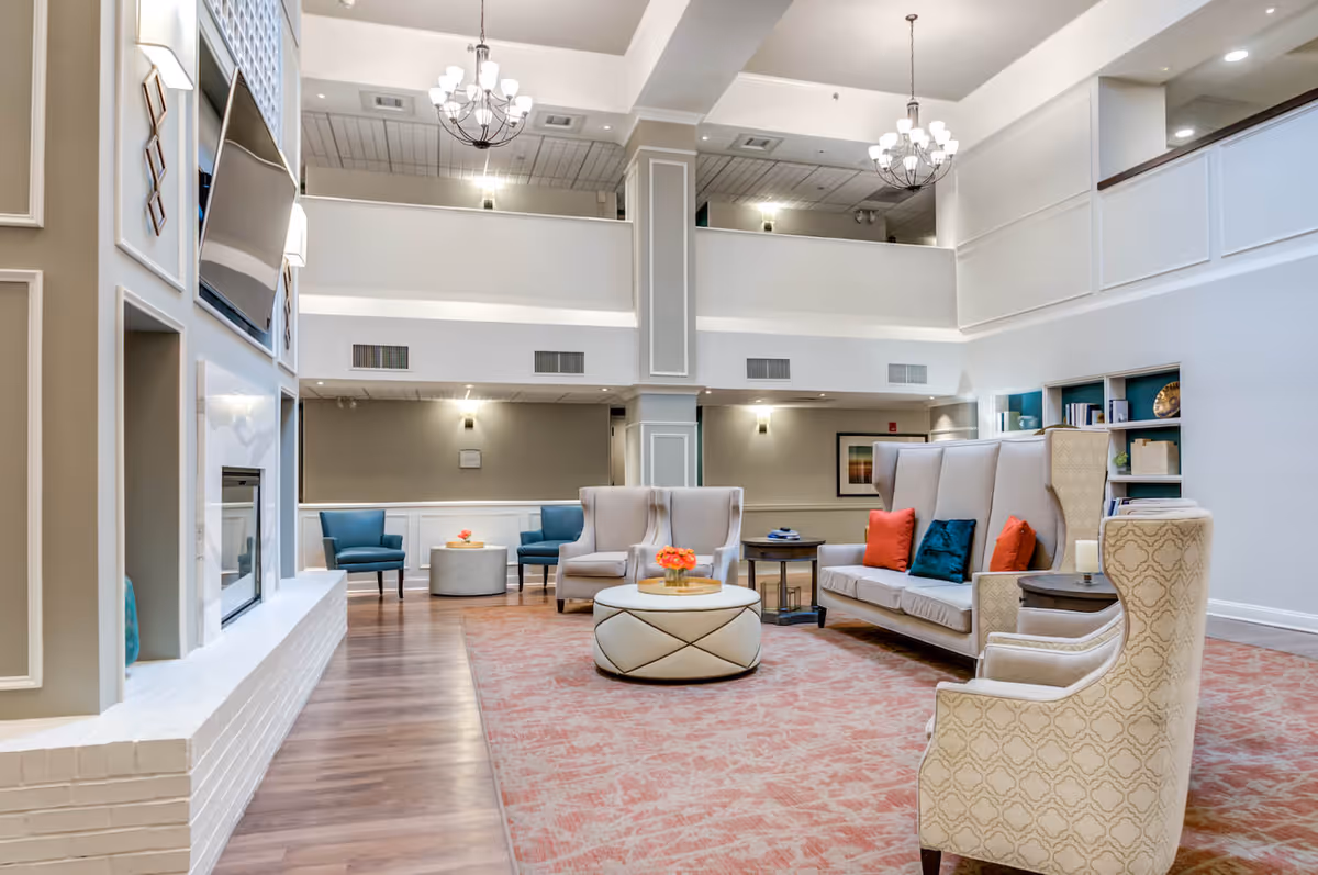 A spacious and elegant living room area with high ceilings, two chandeliers, and a large flat-screen TV mounted above a white brick fireplace. The room features a mix of beige and blue upholstered chairs, a beige sofa with colorful throw pillows, a round ottoman with a flower arrangement, and a patterned area rug on wooden flooring. Built-in shelves with books and decorative items are visible in the background.