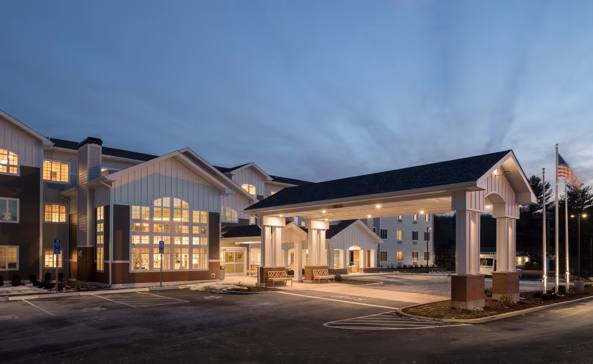 Exterior view of a senior living facility at dusk with a covered entrance, benches, and American flags. The building has multiple windows lit from inside and a parking area in front.