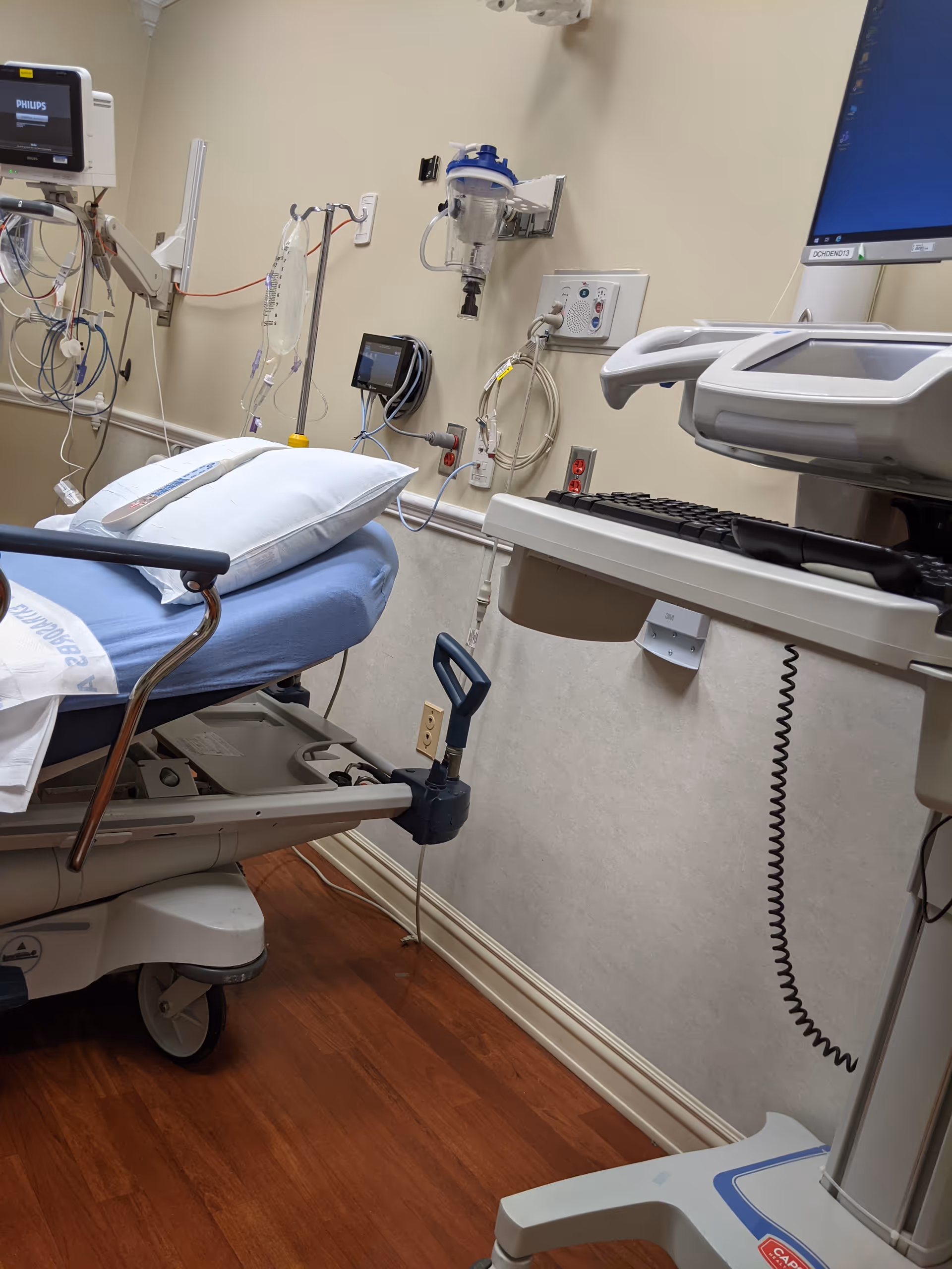 Hospital bed with monitors, IV lines, and computer in a clinical room.