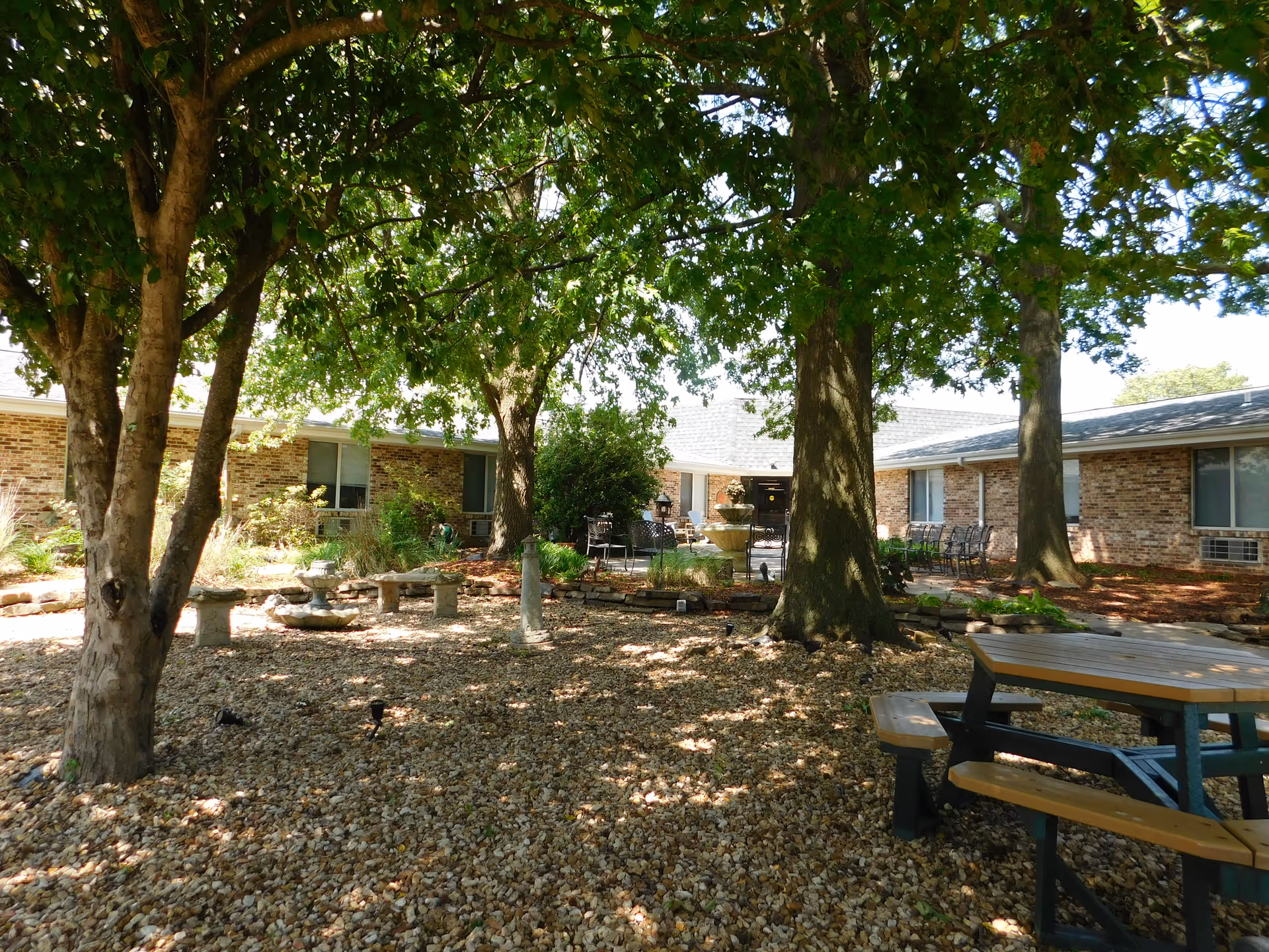 Outdoor courtyard area at Springfield Rehabilitation & HealthCare Center with large trees providing shade, a gravel ground cover, a wooden picnic table with benches, stone benches, a water fountain, and brick building walls with windows in the background.